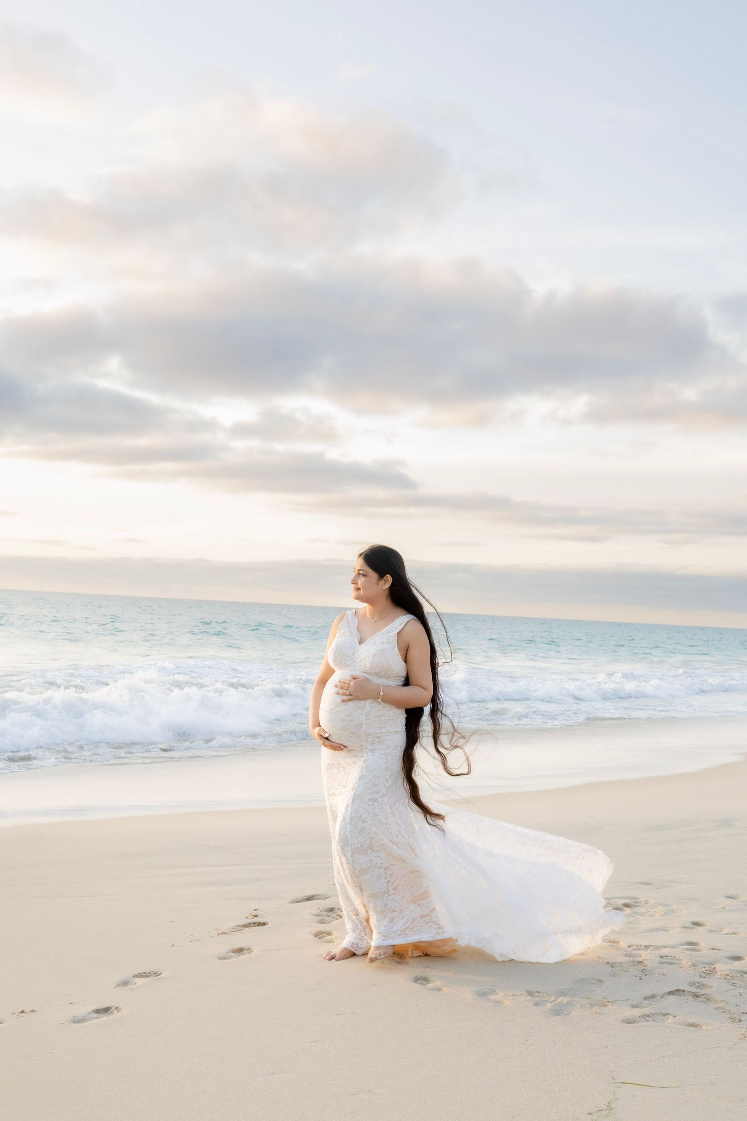Pregnant woman in flowing white lace gown cradling belly on serene beach at sunset.