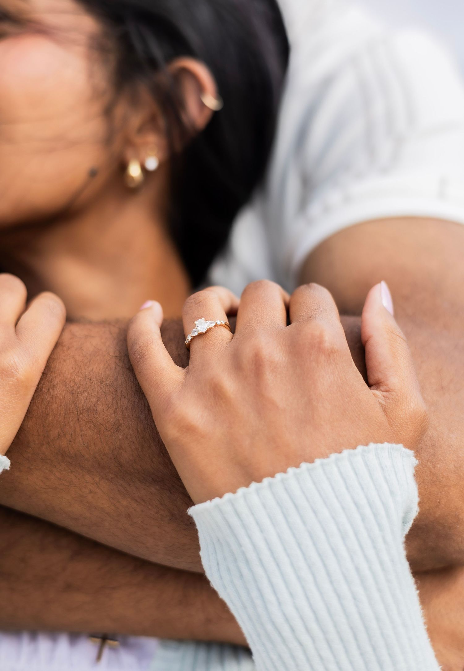 Close-up of womans hand with diamond engagement ring wrapped around partners arm outdoors.