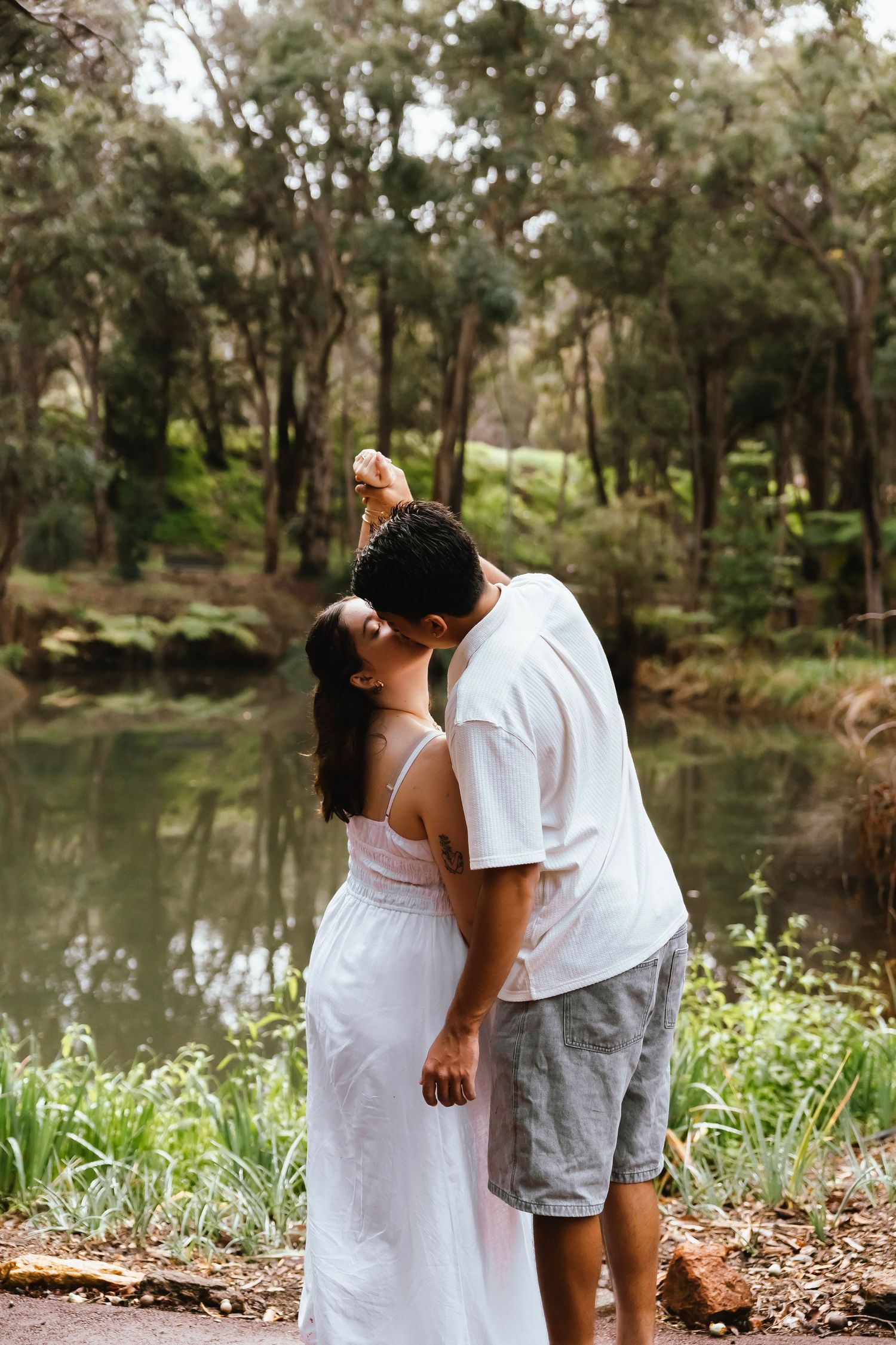 Couple sharing a romantic kiss by a serene pond surrounded by tall eucalyptus trees in nature.