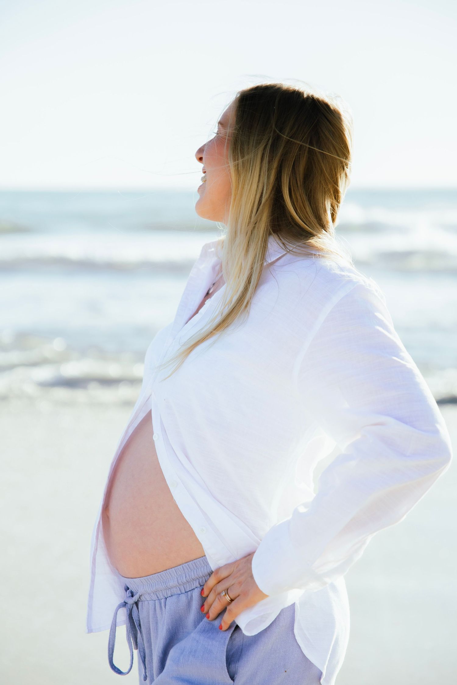 Pregnant woman in white shirt and blue pants smiling at the ocean on a beach.
