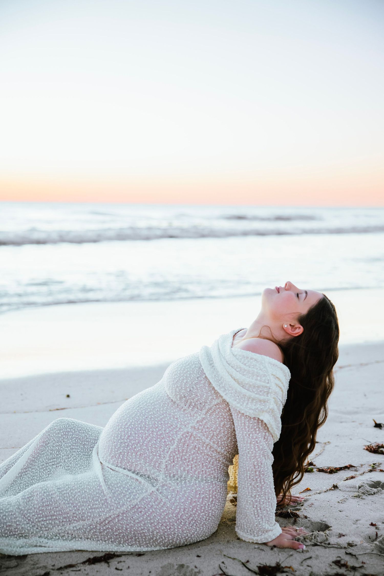 Pregnant woman in white knit dress reclines on beach at sunset, head tilted back with long dark hair flowing.