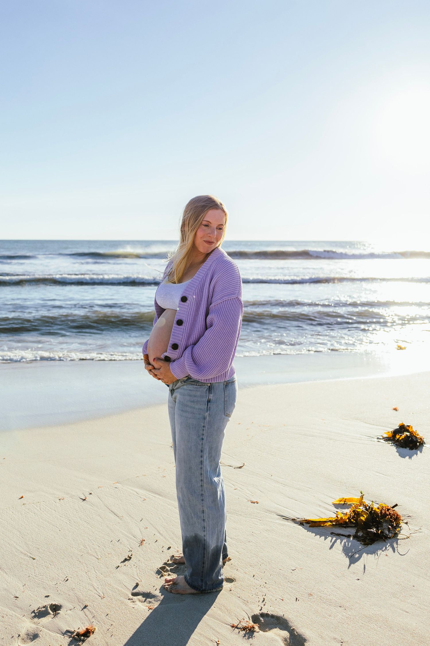 Smiling pregnant woman in purple cardigan and jeans poses on sunny beach with ocean backdrop.