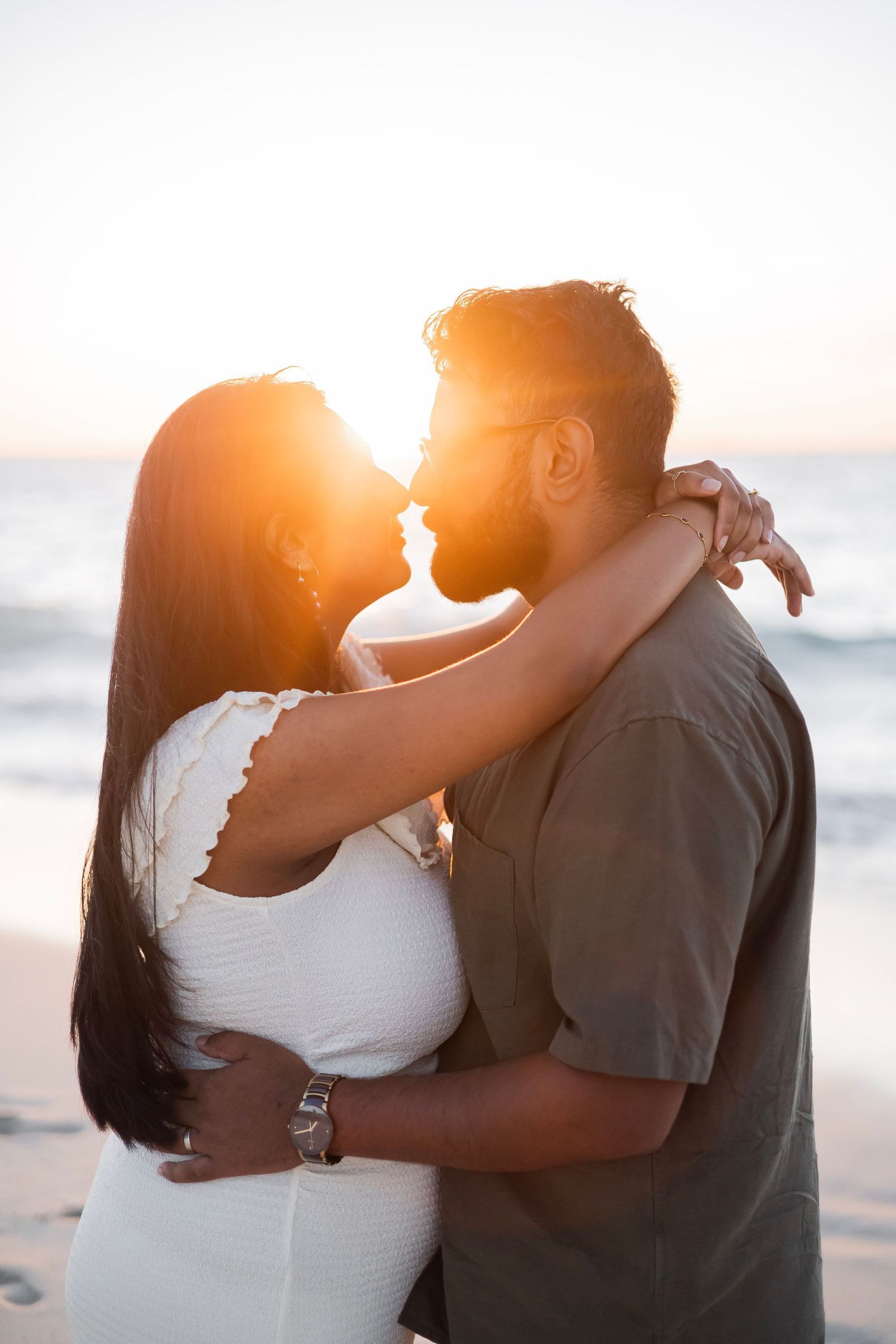 Couple embracing and kissing on a sunny beach at sunset, backlit by warm golden light near the ocean.