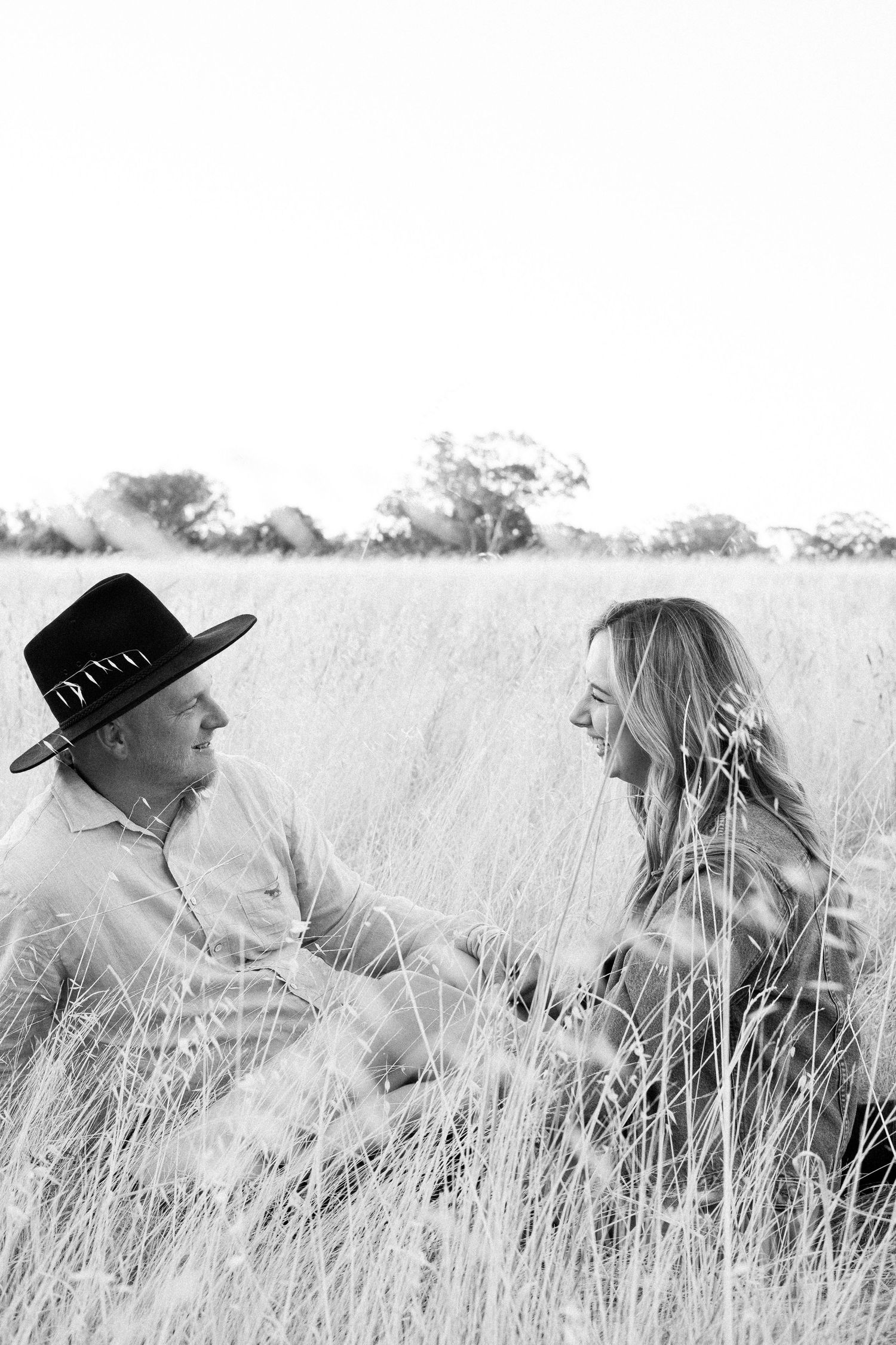 Black and white photo of couple sitting and laughing together in tall golden grass field.