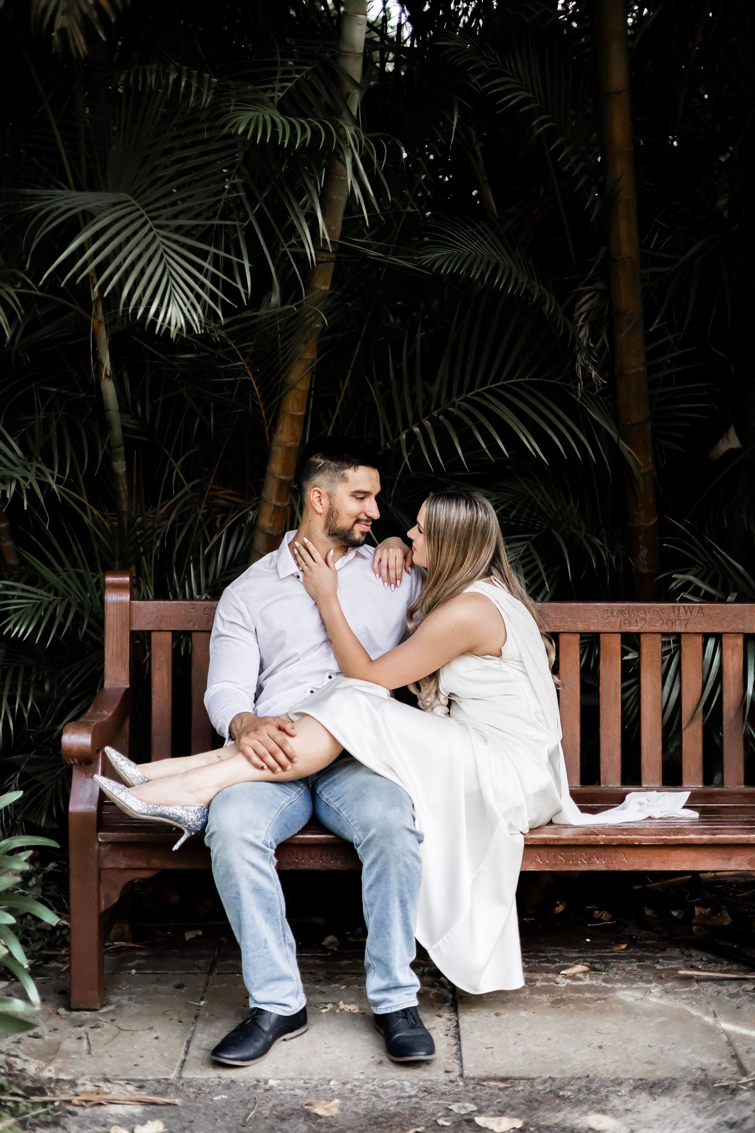 Couple sitting closely on a wooden bench, sharing an intimate moment amid lush tropical palm tree backdrop.