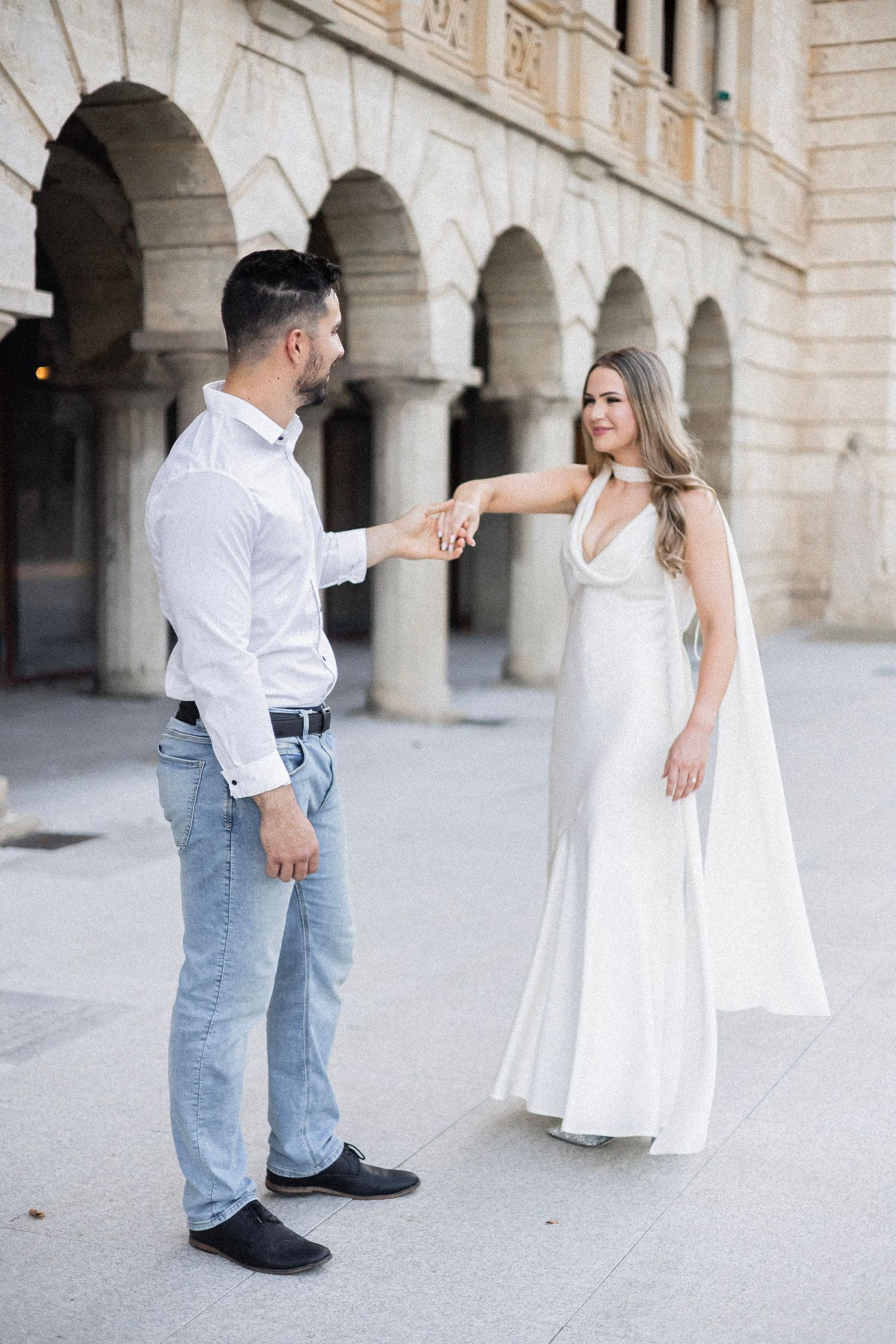 Engaged couple holding hands facing each other near stone archway colonnade during engagement session.