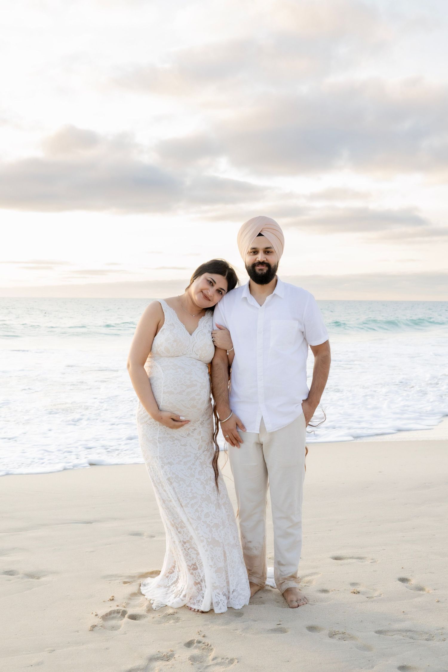 Maternity beach portrait of couple in white outfits posing on sandy shore at sunset.
