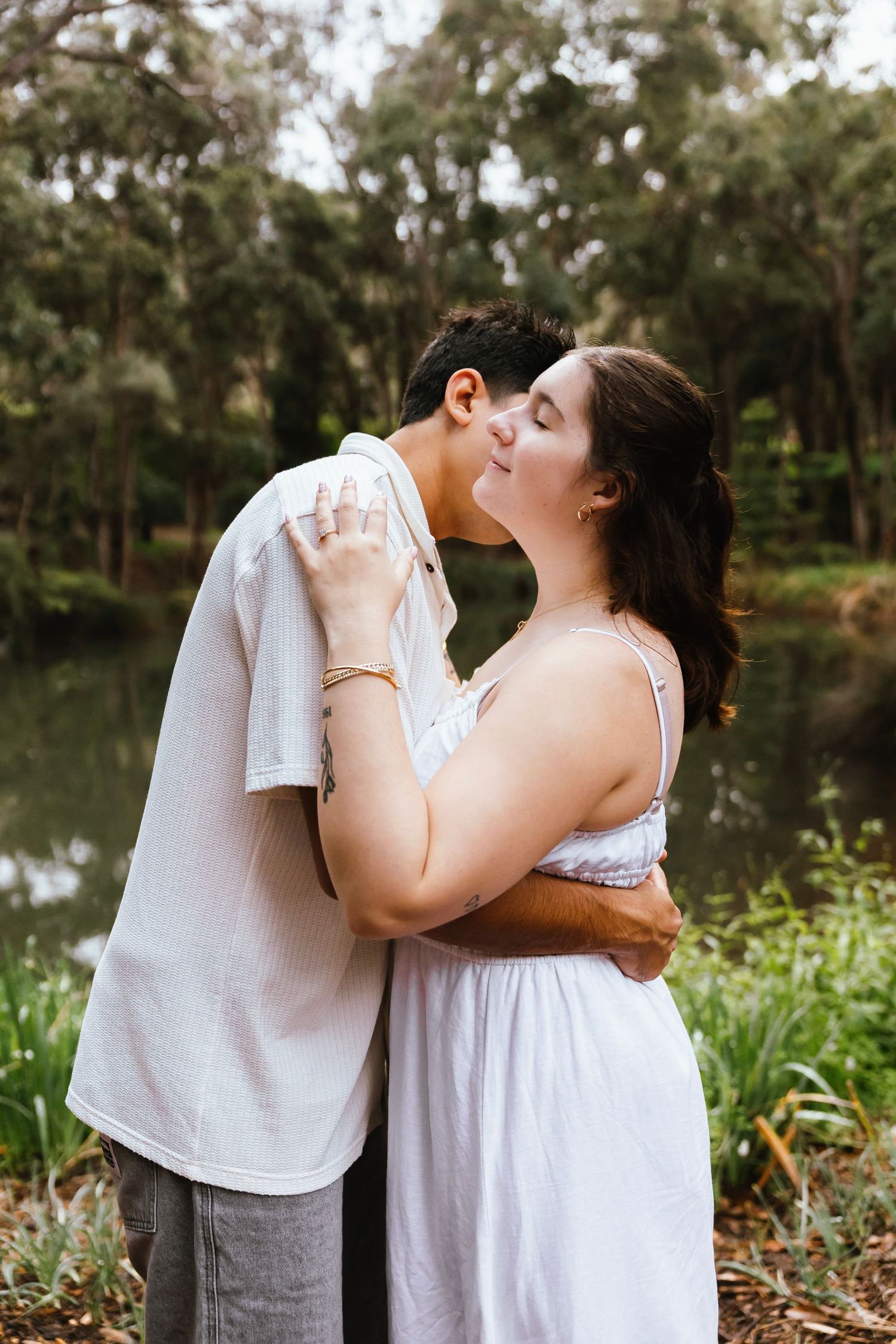 Romantic couple sharing a kiss near a tranquil pond surrounded by lush green foliage.