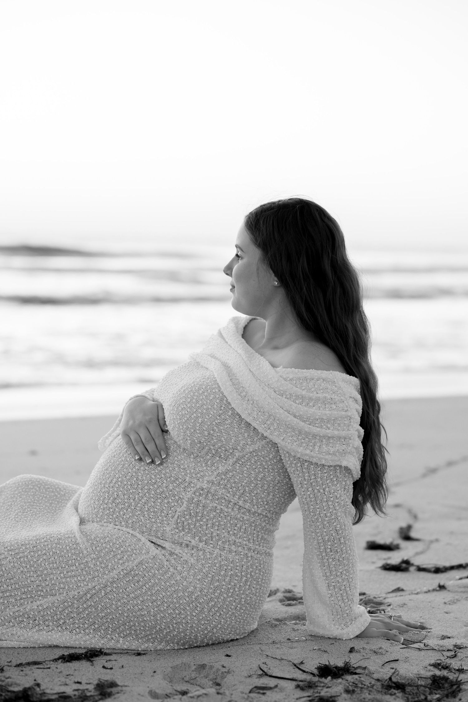 Pregnant woman in white lace dress sitting on beach sand, gazing at ocean waves in black and white photo.