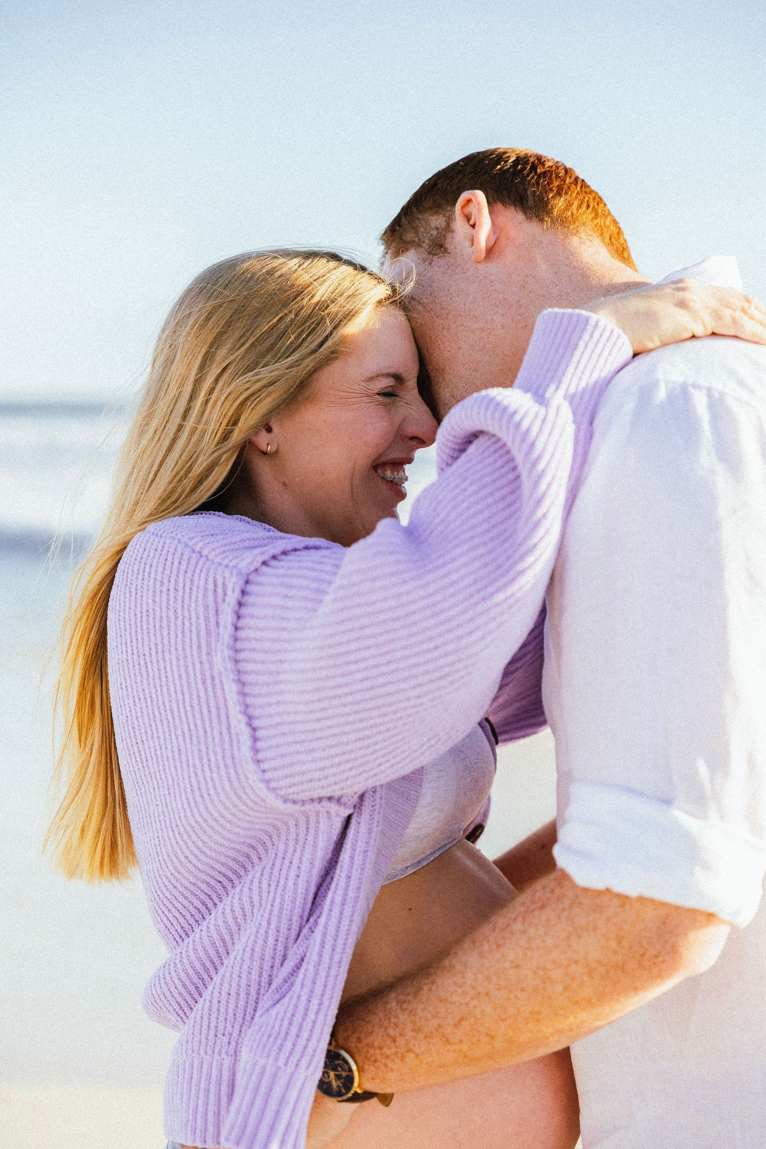 A happy couple embraces and laughs together on a sunny beach, woman in lavender sweater, man in white shirt.