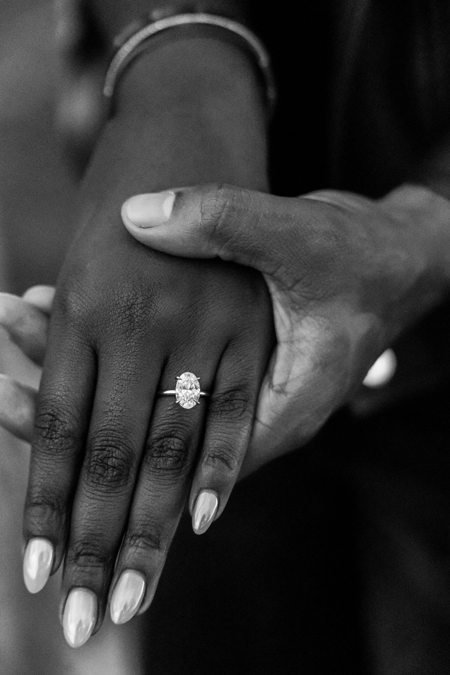 Black and white photo of hands displaying a large oval diamond engagement ring with manicured nails and a bracelet.