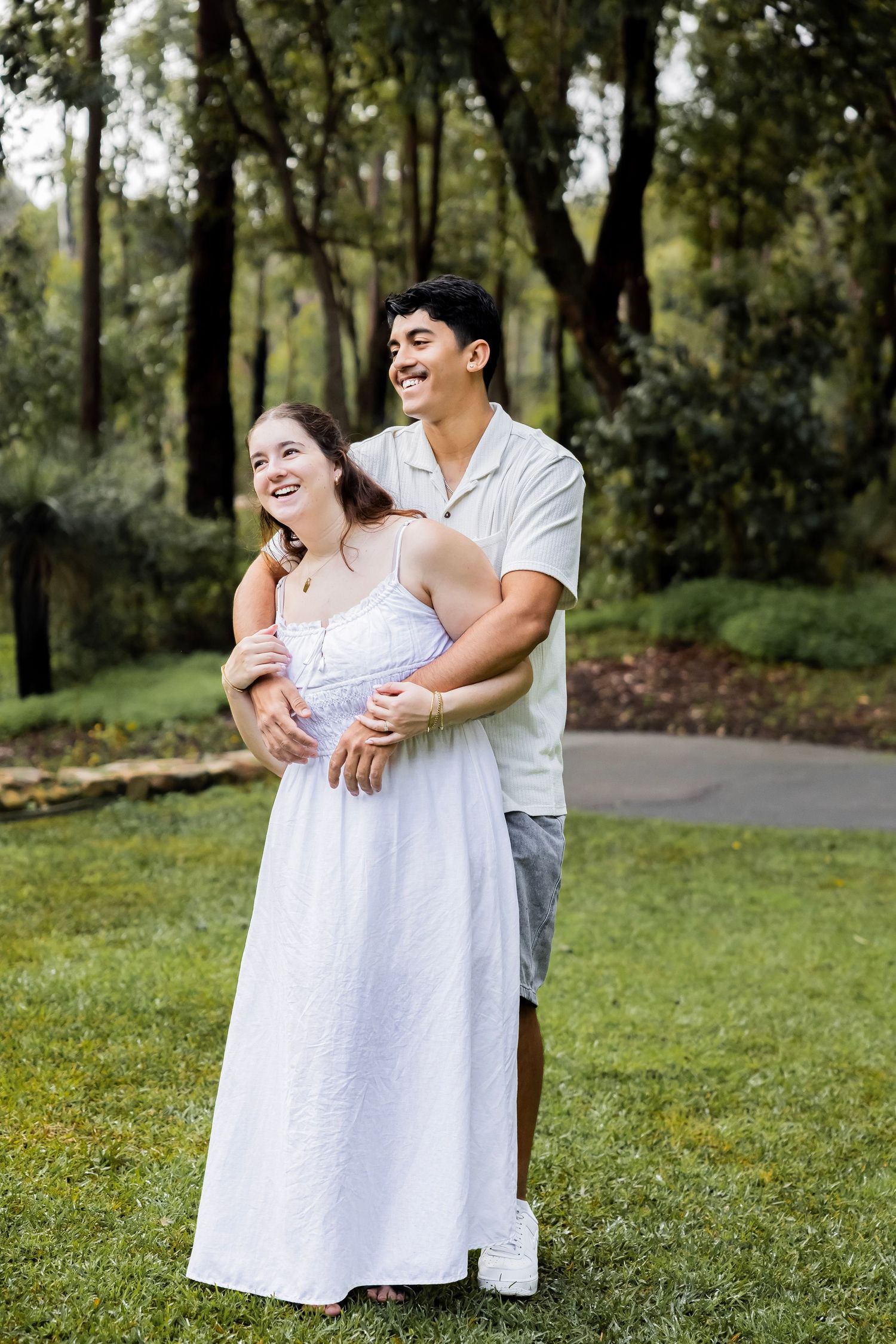 Happy couple embracing and laughing together on a green lawn surrounded by tall trees.