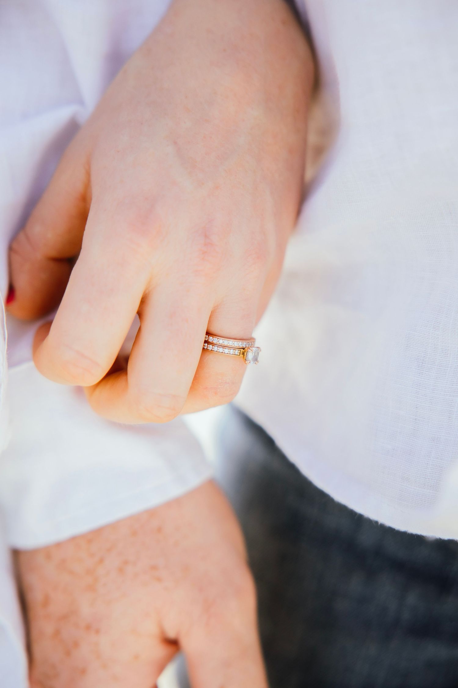 Close-up of a hand wearing a delicate gold diamond ring, held against a white shirt.