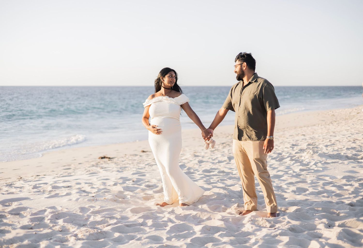 Pregnant couple holding hands on a sunny white sand beach, woman in white maternity gown, man in olive shirt.