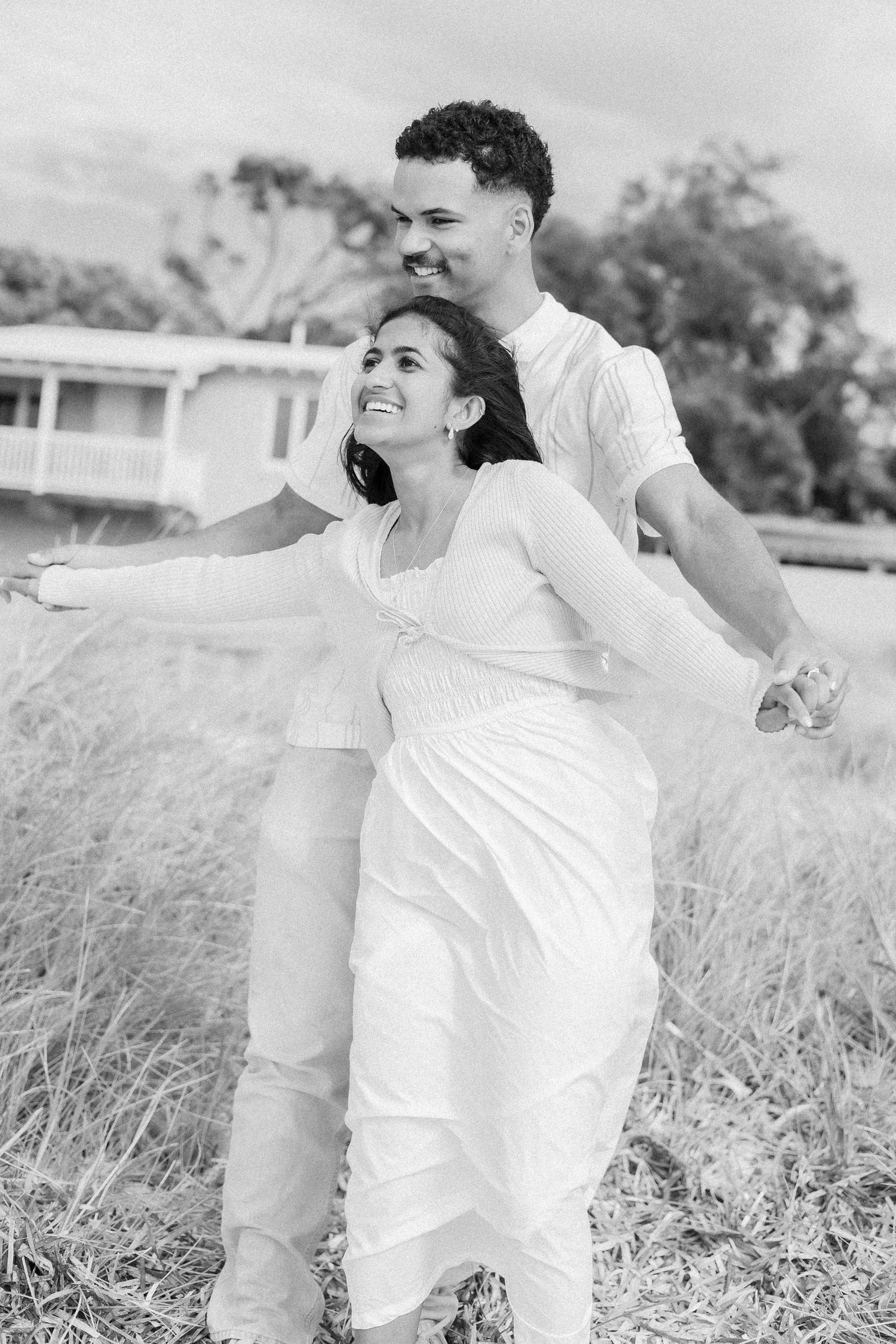 Black and white photo of joyful woman with arms outstretched, man smiling behind her in field.