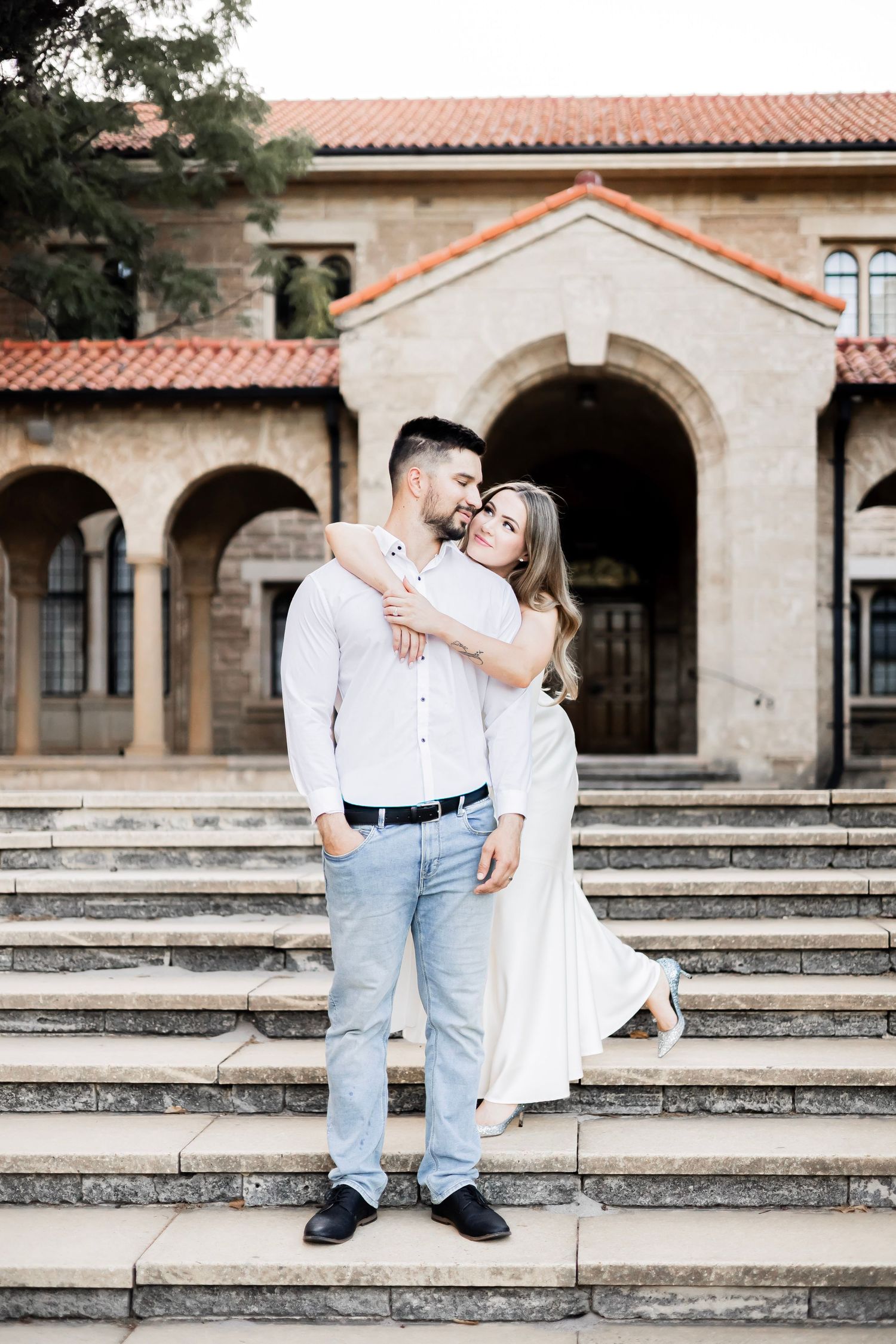 Man and woman in white attire sharing a tender embrace on steps outside a stone archway building.