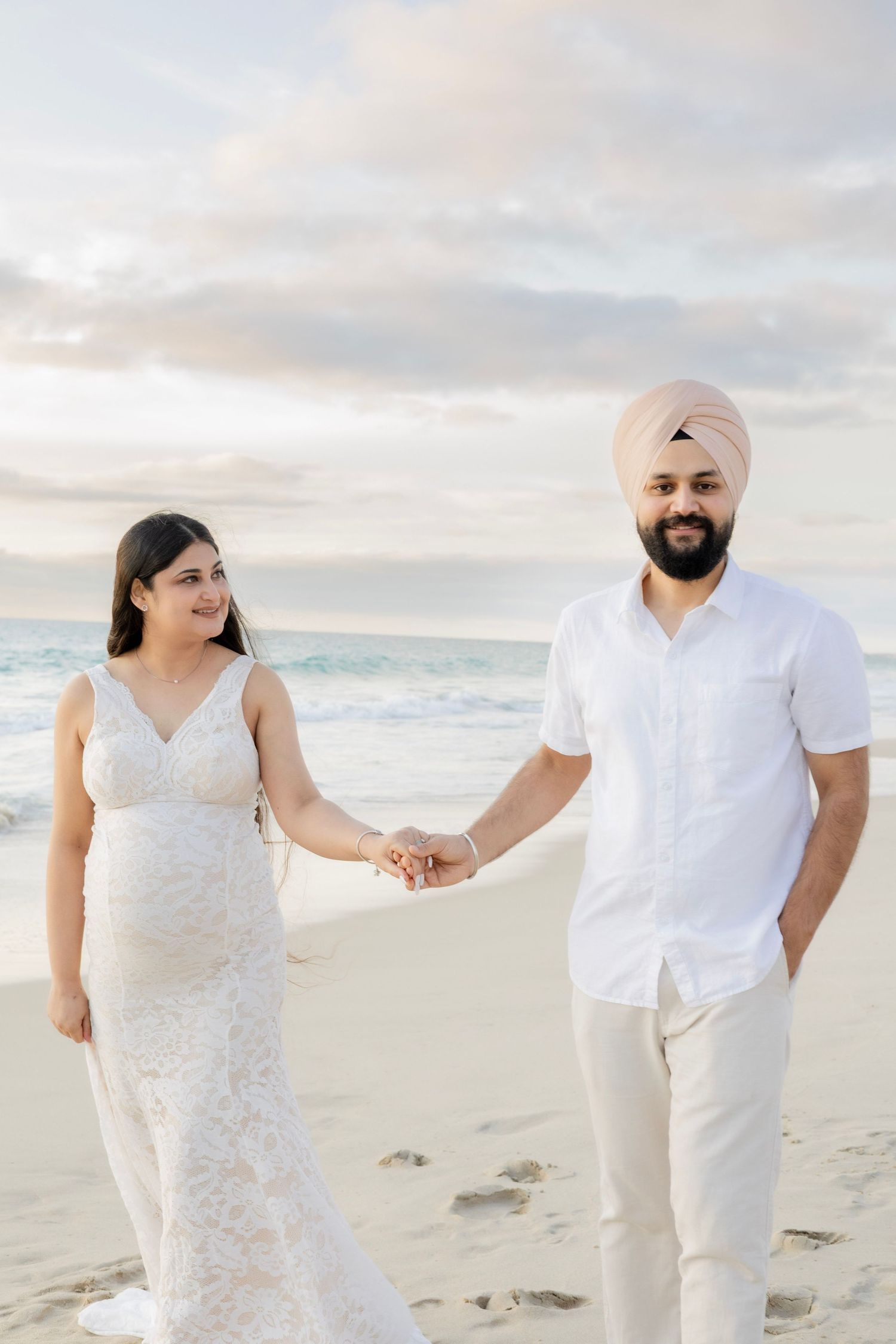 Pregnant couple holding hands and smiling on beach at sunset, man wearing pink turban and white shirt.