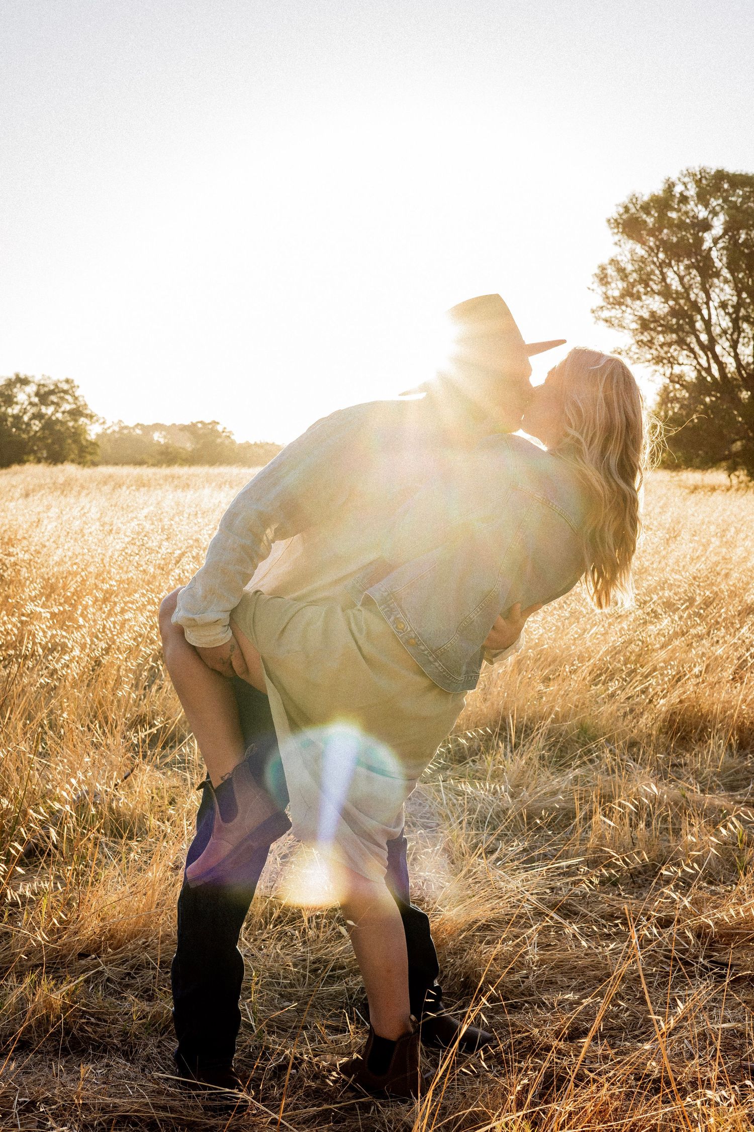 A couple shares a romantic lift and kiss in a field of golden grass with bright sun flare.