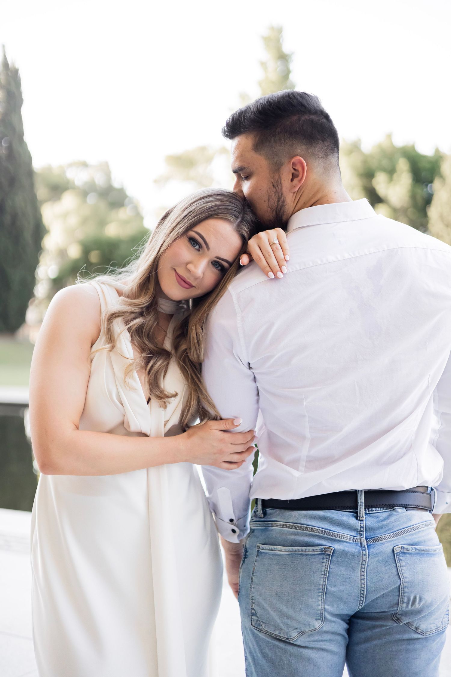 Couple embracing tenderly outdoors, woman in white dress resting head on mans shoulder near pond.