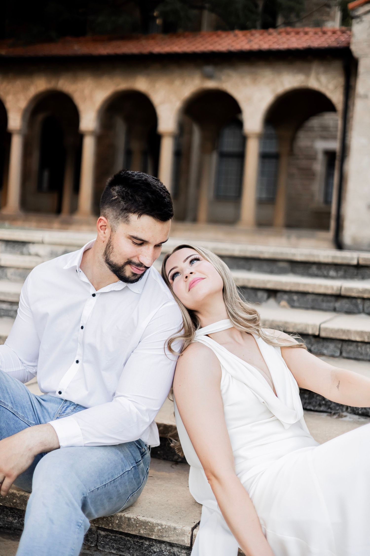 Romantic couple sitting on stone steps, woman in white halter dress leaning back on man in white shirt and jeans outdoors.