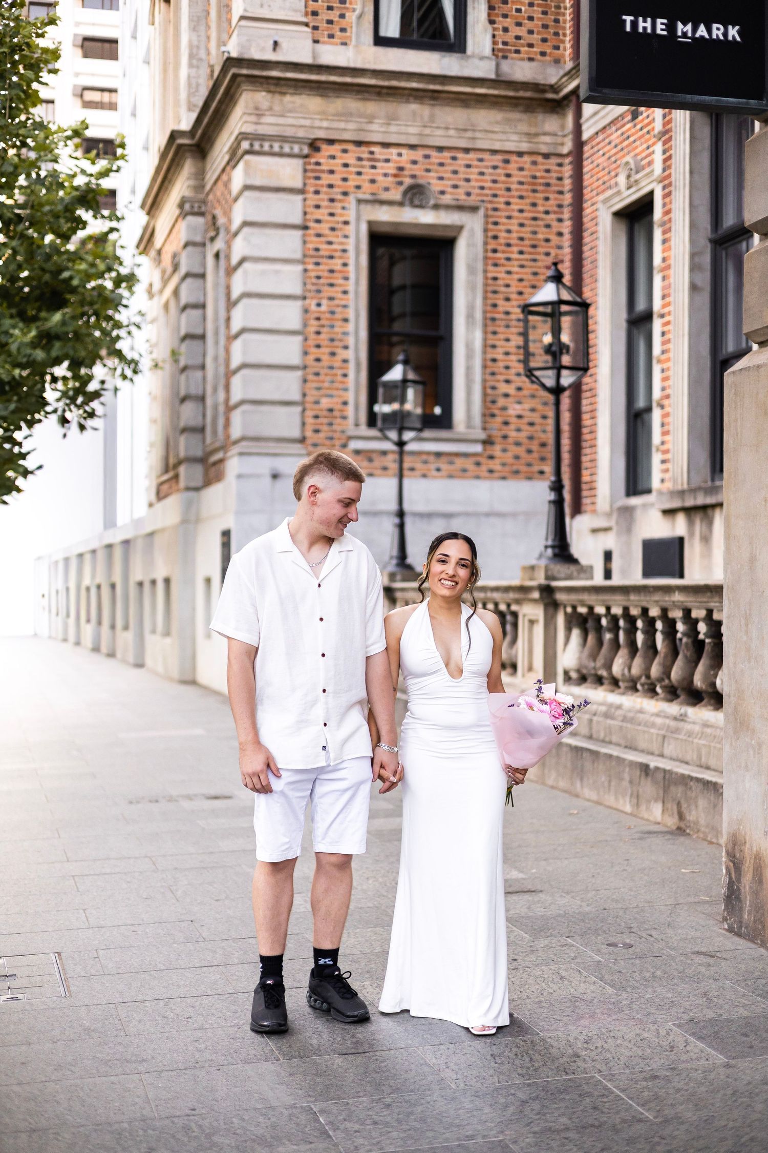 Smiling couple in white holding hands and flowers outside The Mark building.