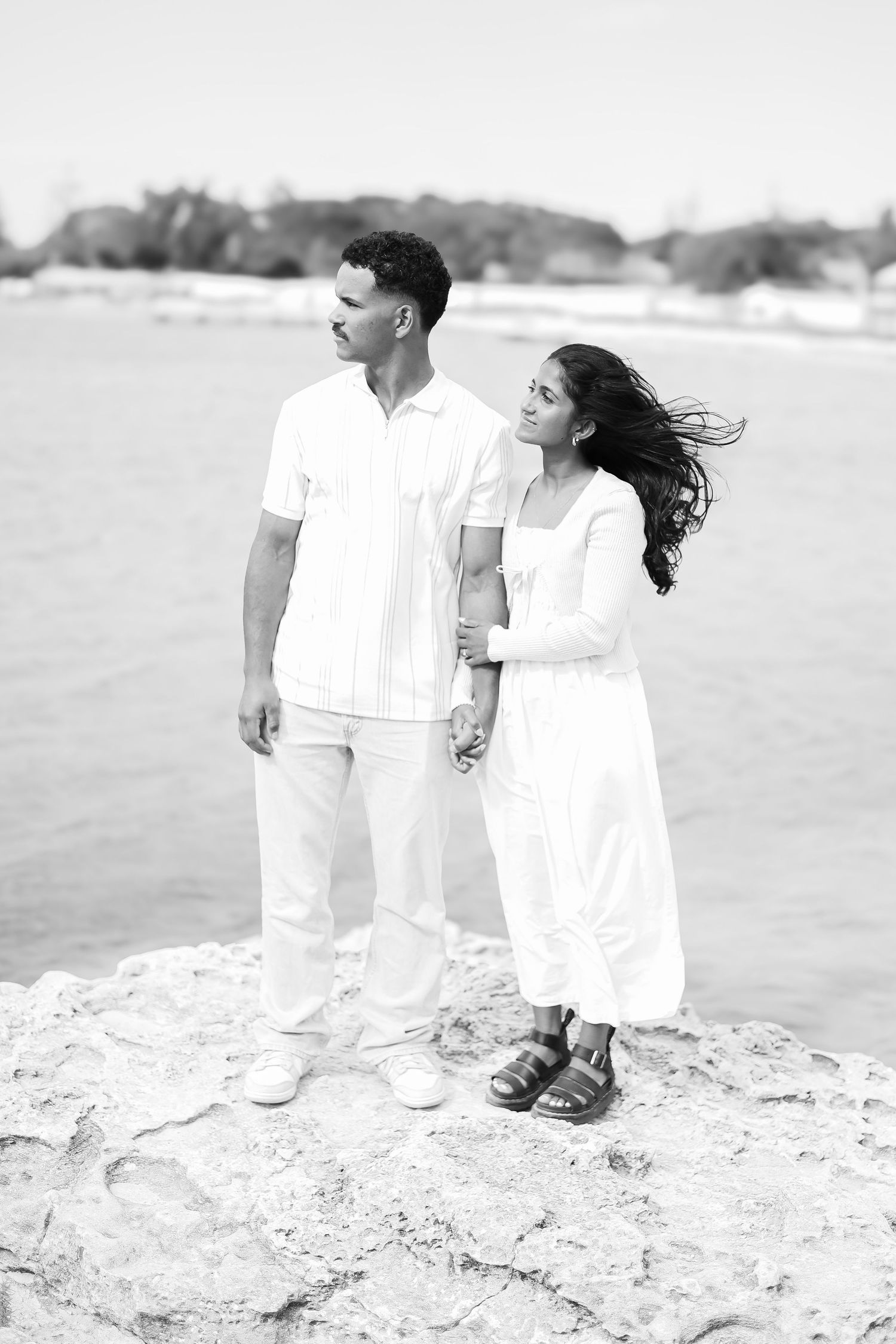 Black and white photo of a couple in white outfits standing on rocks by the ocean, woman resting her head on mans shoulder.