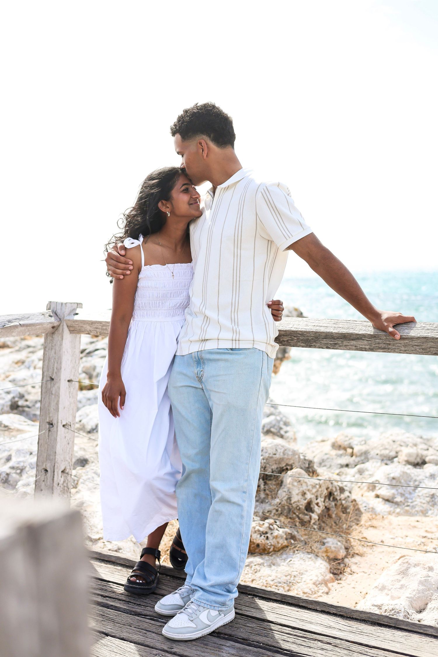 Couple standing on a wooden boardwalk by the ocean, man kissing woman's head affectionately.