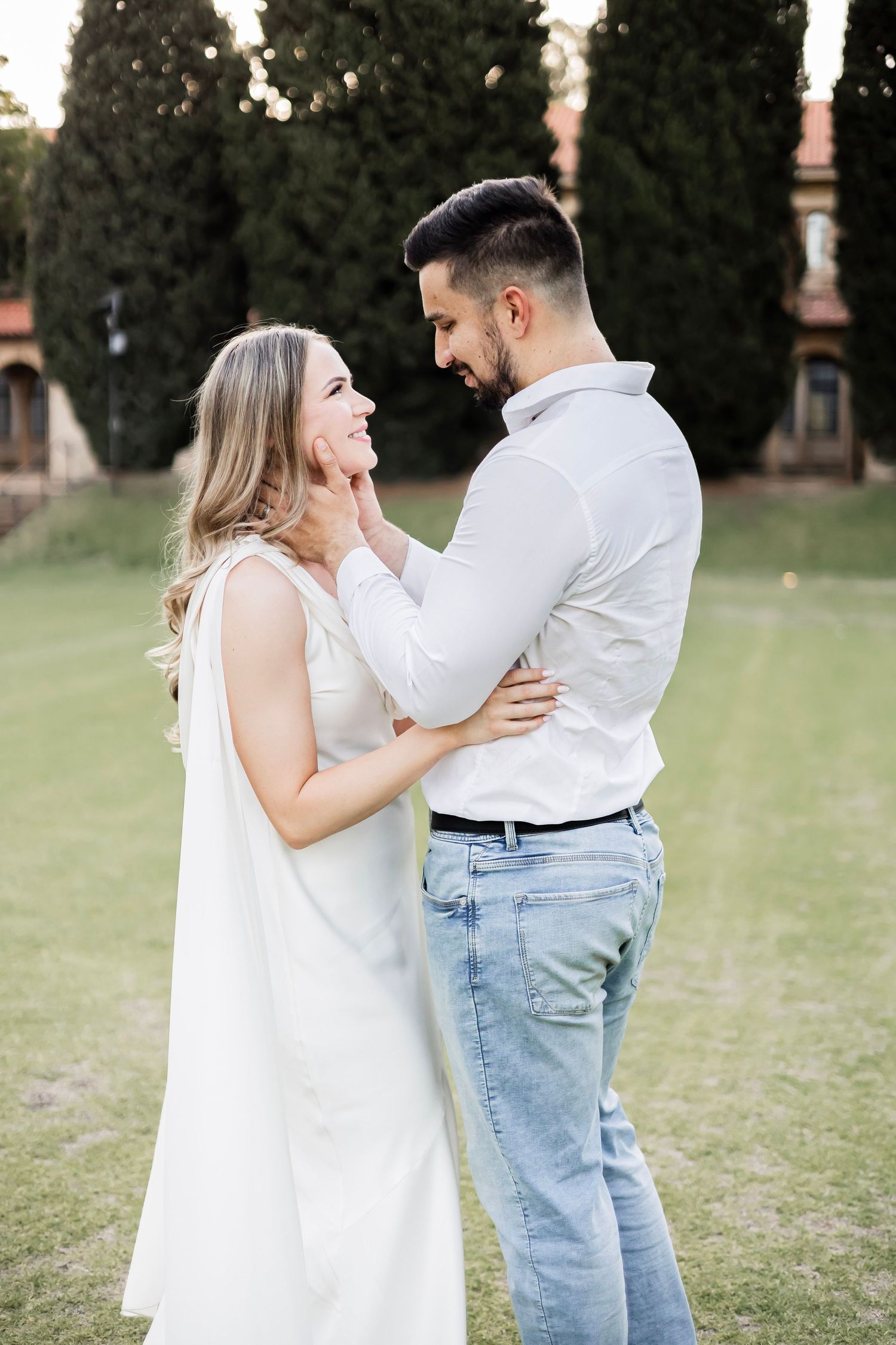 Couple in white outfits share a tender moment on green lawn with cypress trees and historic building behind.