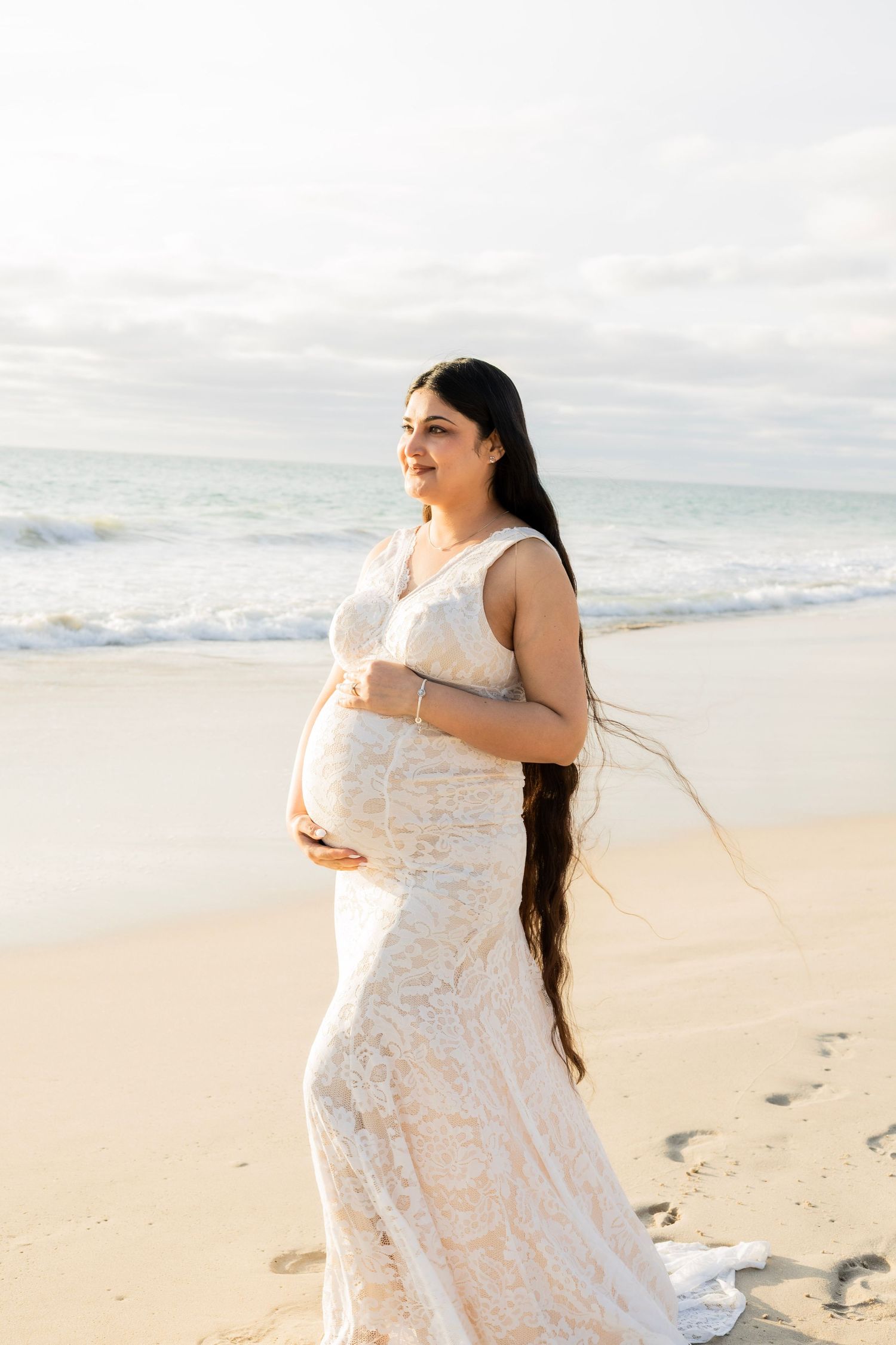 Pregnant woman in white lace gown cradling baby bump on beach, long hair flowing in the breeze.