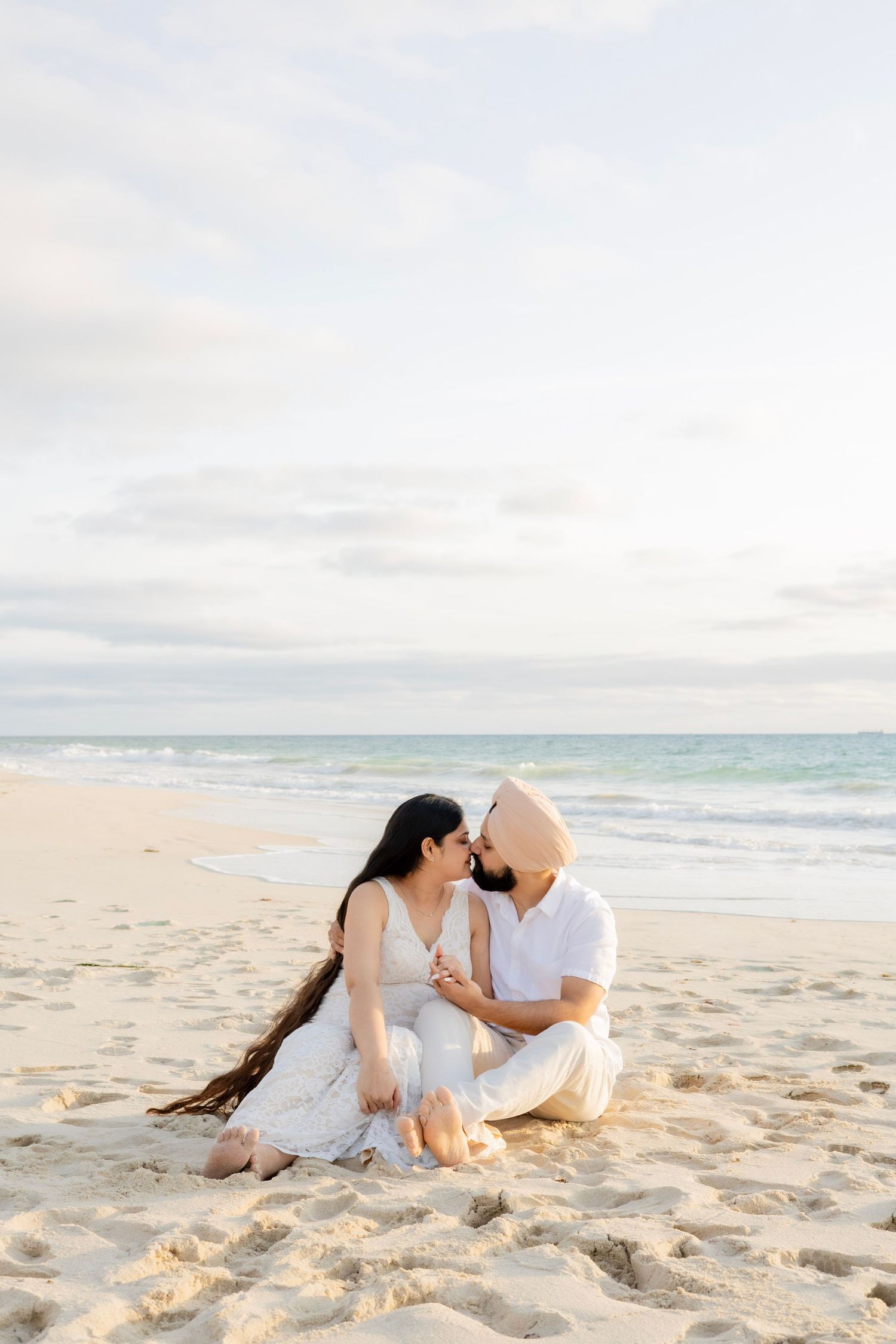Couple sitting on sandy beach at sunset, sharing a romantic moment with ocean waves in the background.
