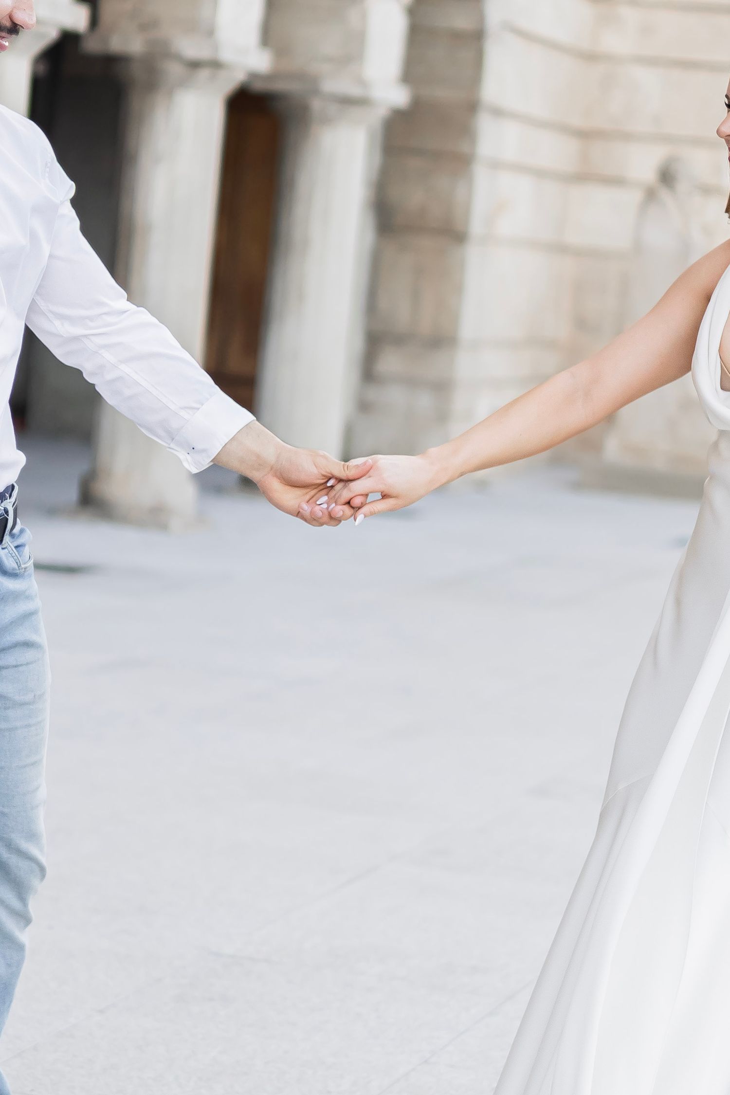 A couple holds hands outdoors near elegant stone columns, with the bride in a white gown and groom in jeans.