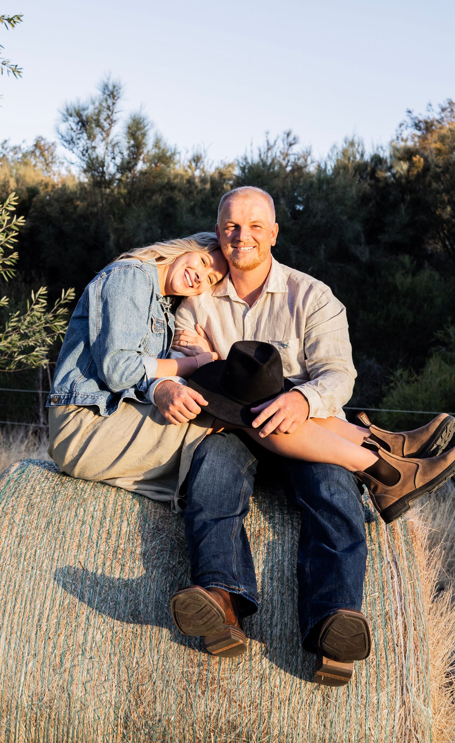 Couple sitting on a hay bale smiling, man holding cowboy hat, woman leaning on his shoulder, rural setting.