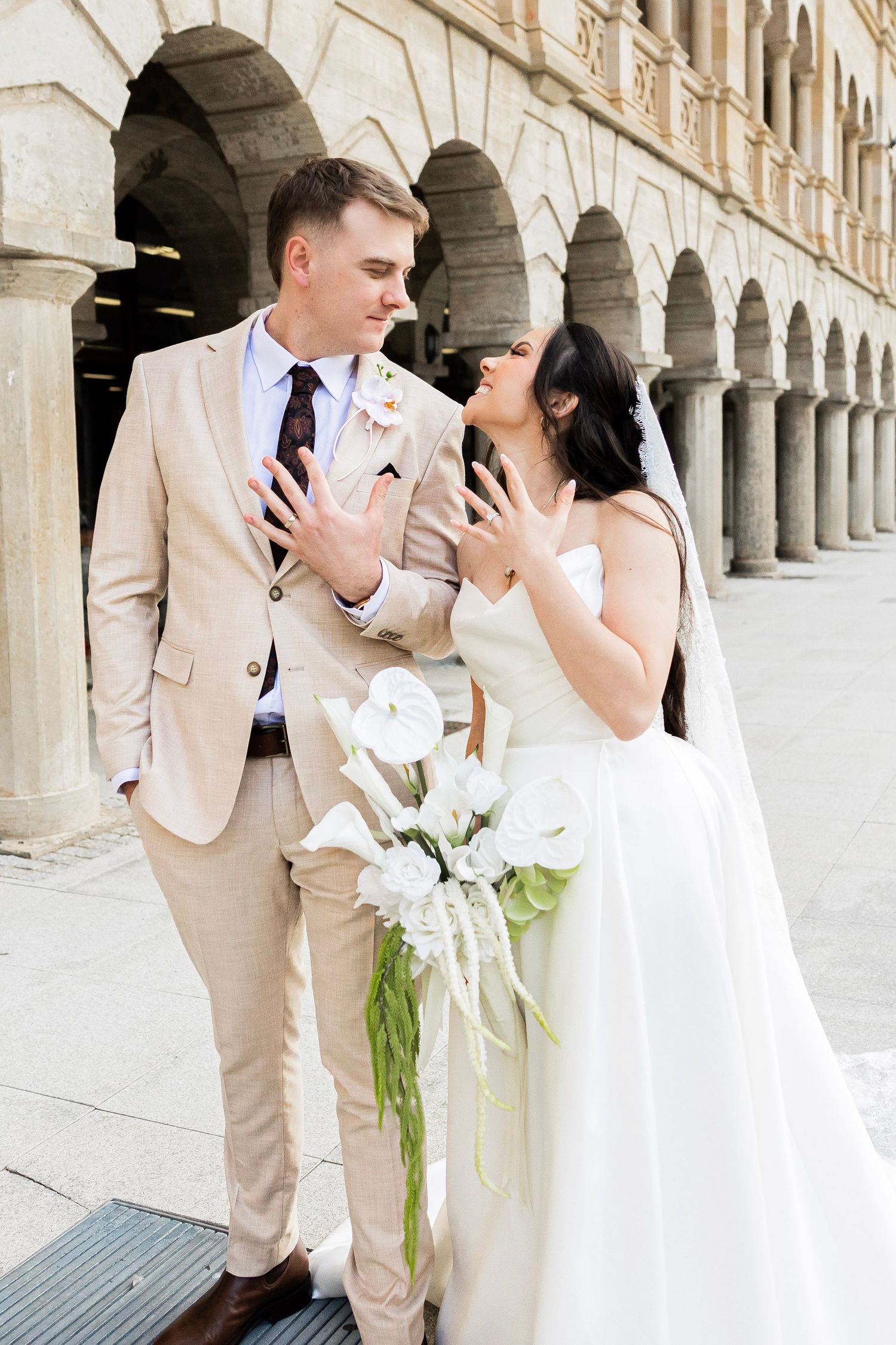 Bride and groom share a joyful moment outside historic arched building, bride holding white calla lily bouquet.