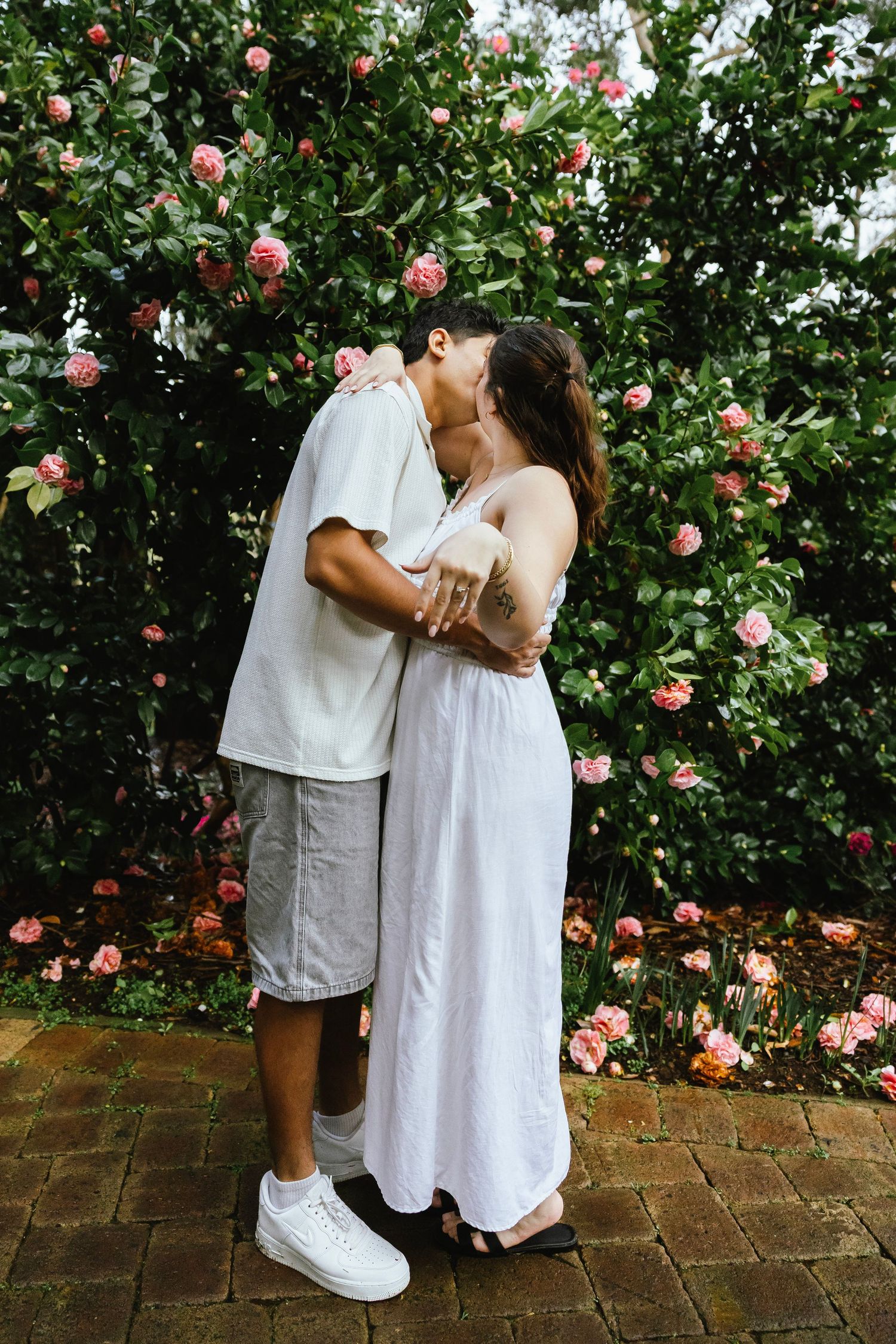 Engaged couple kissing while embracing in front of a lush wall of pink roses.