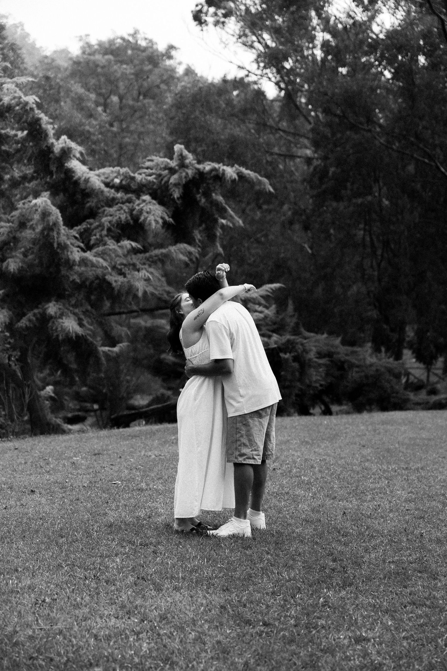 Black and white photo of a couple embracing and dancing together on a lawn surrounded by trees in a park.