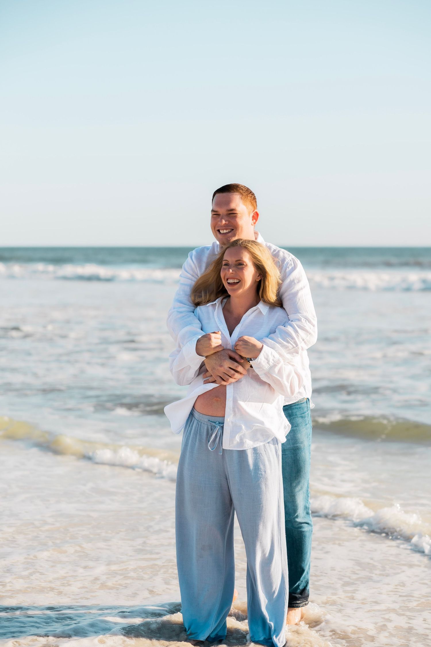 Happy couple embracing on a sunny beach, wearing white shirts and light pants, with ocean waves in the background.