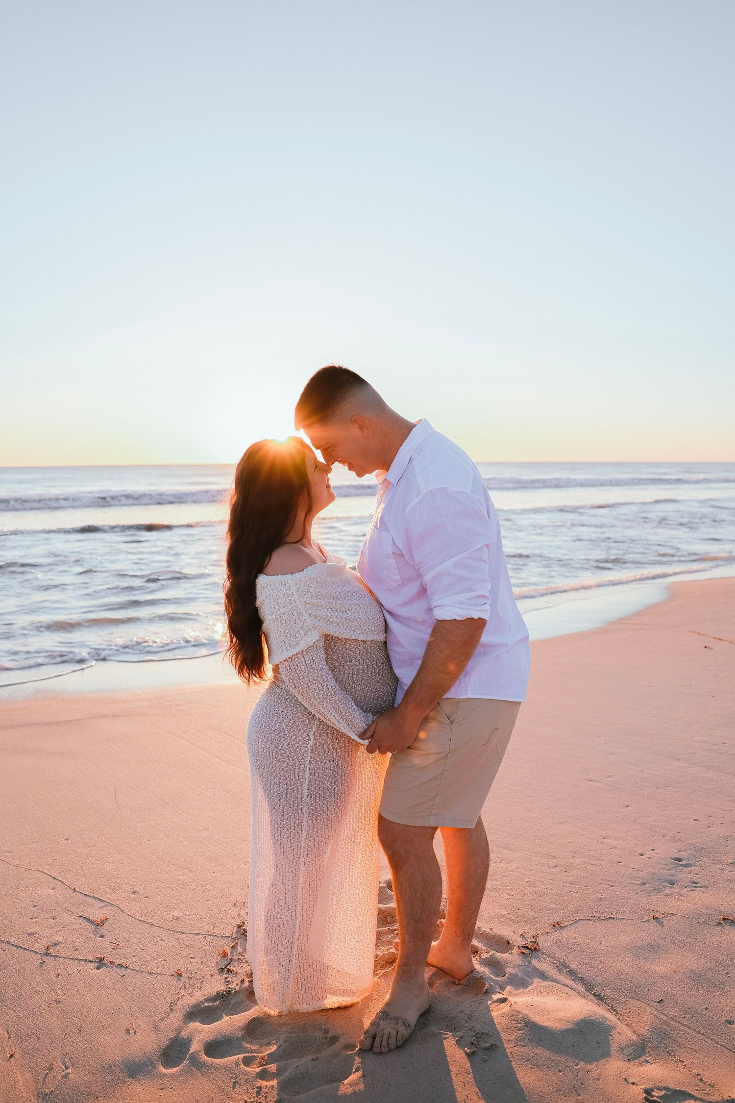 Pregnant couple holding hands on sandy beach at sunset, touching foreheads in tender embrace.