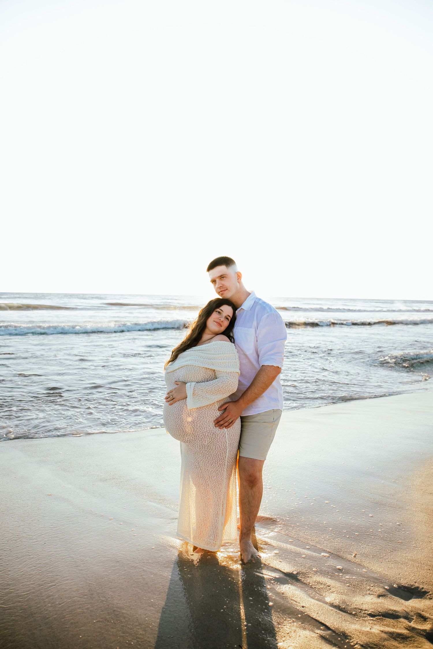 Pregnant couple embracing on a sunny beach, woman in white maxi dress, man in light blue shirt and gray shorts.