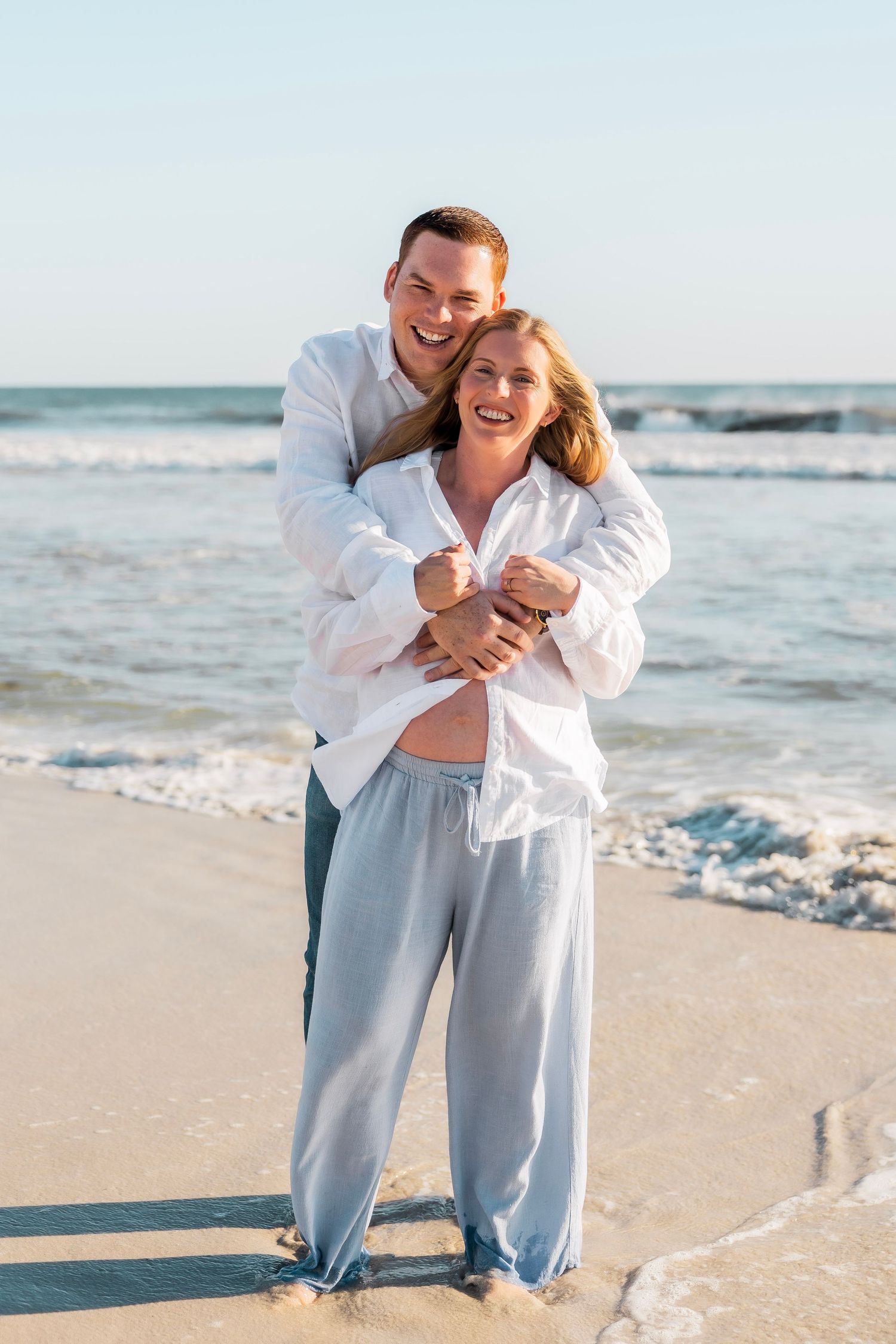 Happy couple embracing on a sunny beach, woman in white shirt and blue pants, waves in background.