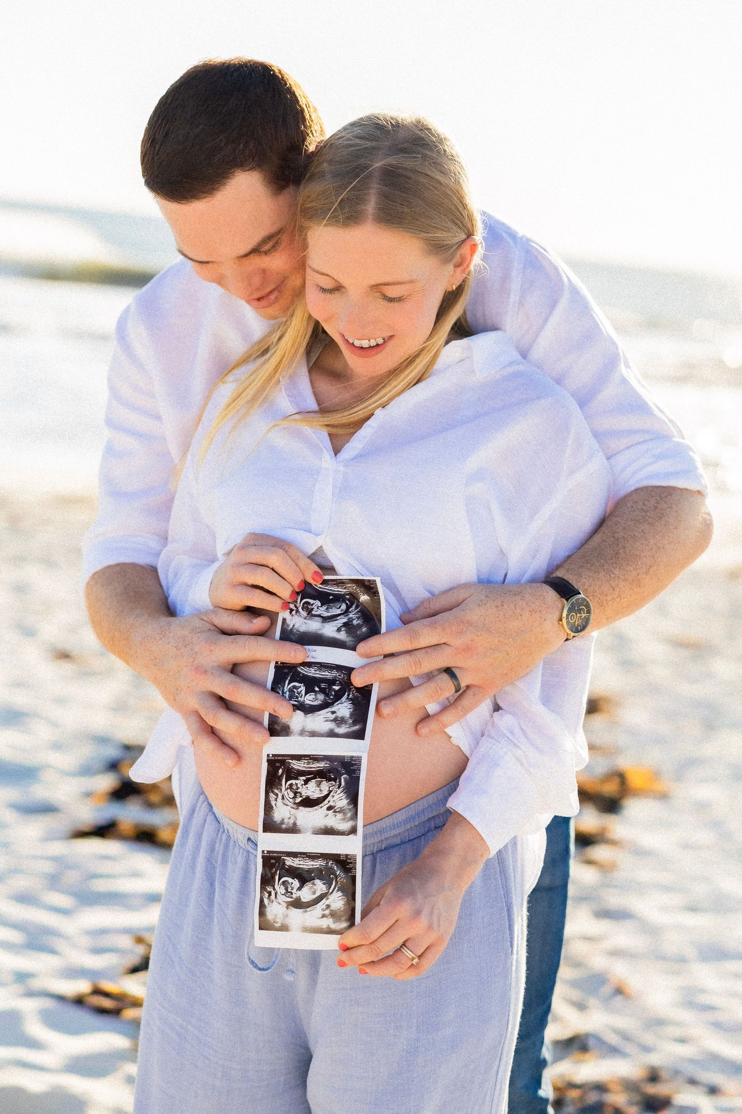 Expecting couple on beach holding ultrasound photos against pregnant belly, smiling joyfully.