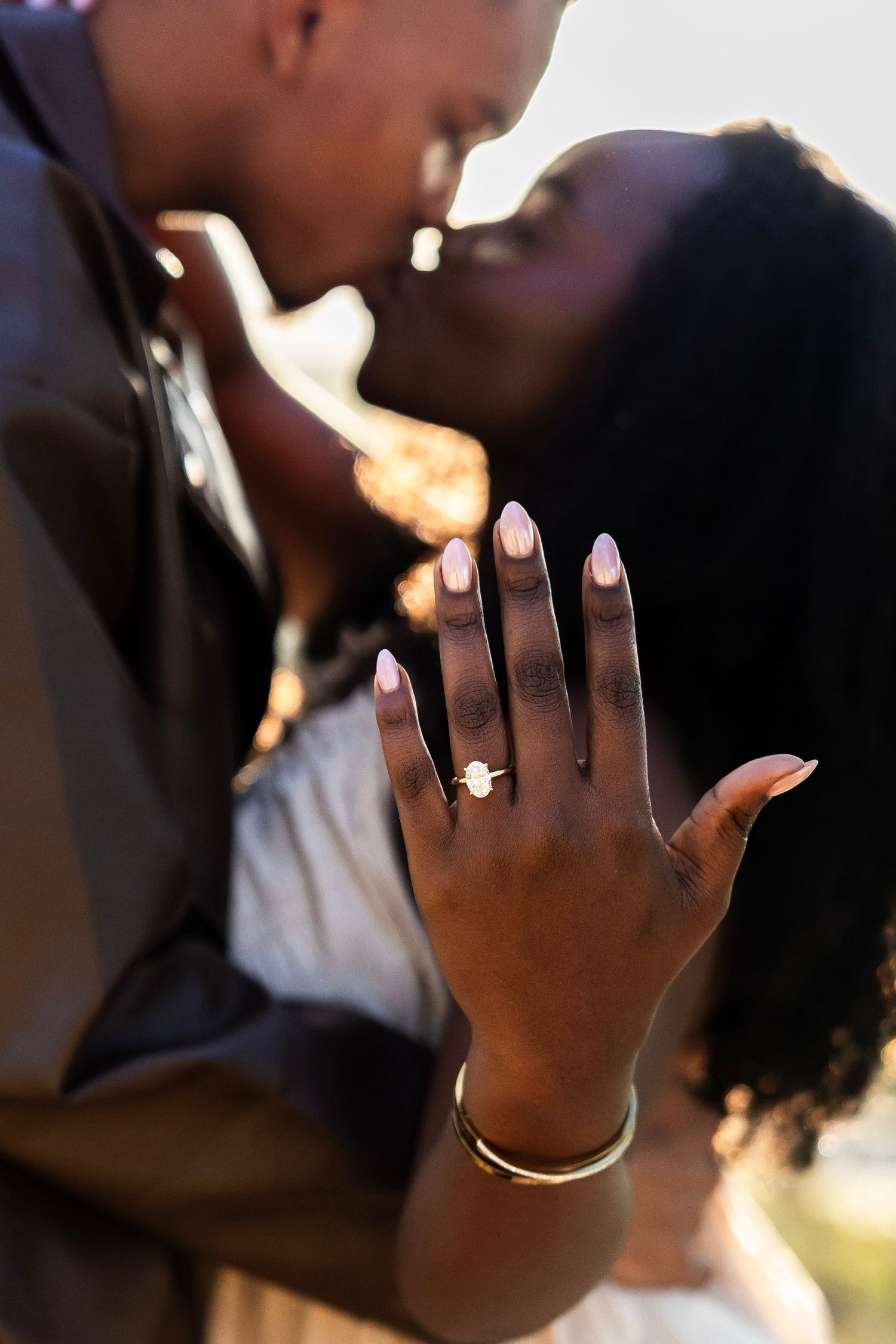 Couple kissing while showing off engagement ring with gold bracelet in warm sunset light.