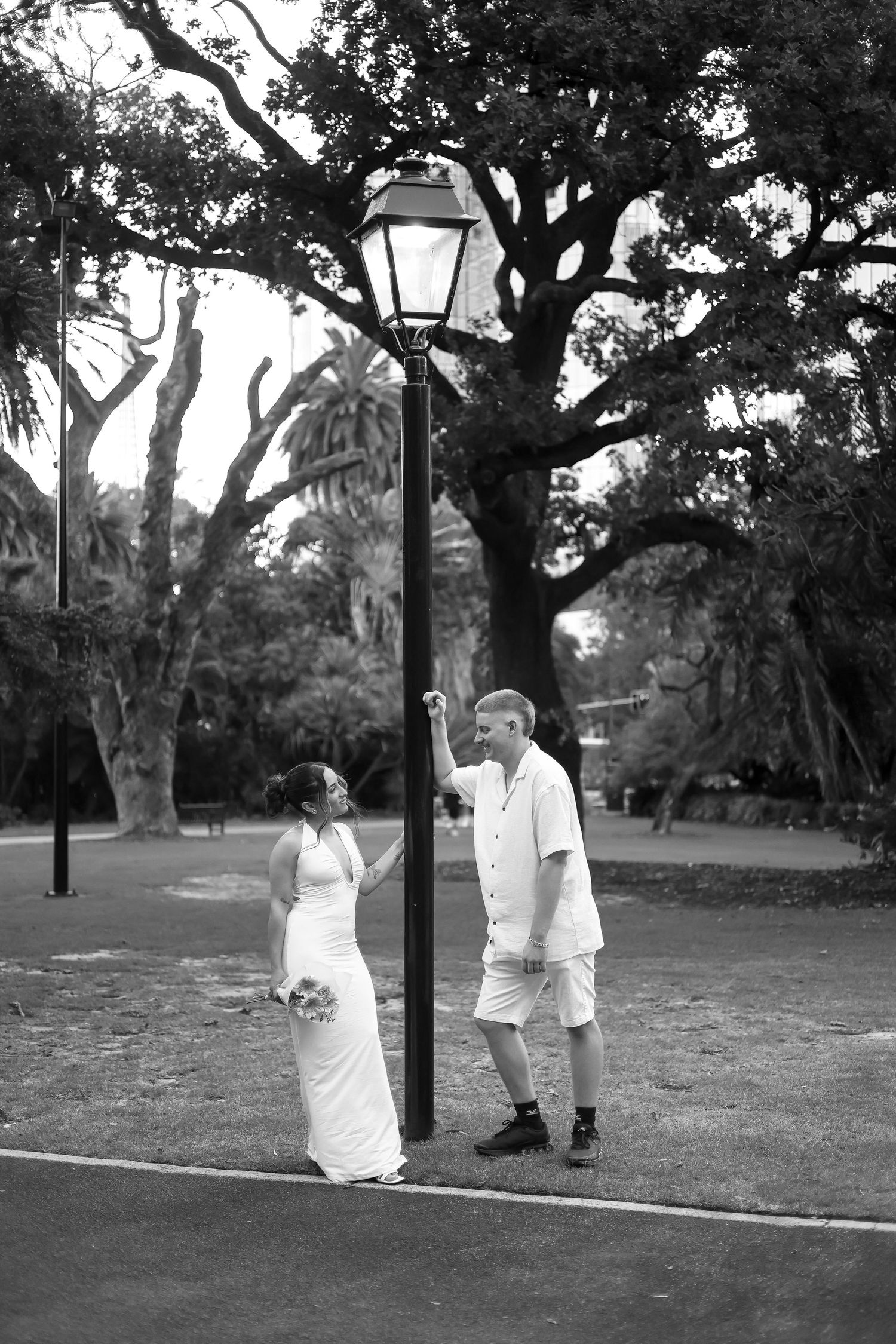 Black and white photo of a bride and groom posing by a vintage street lamp in a lush park setting.