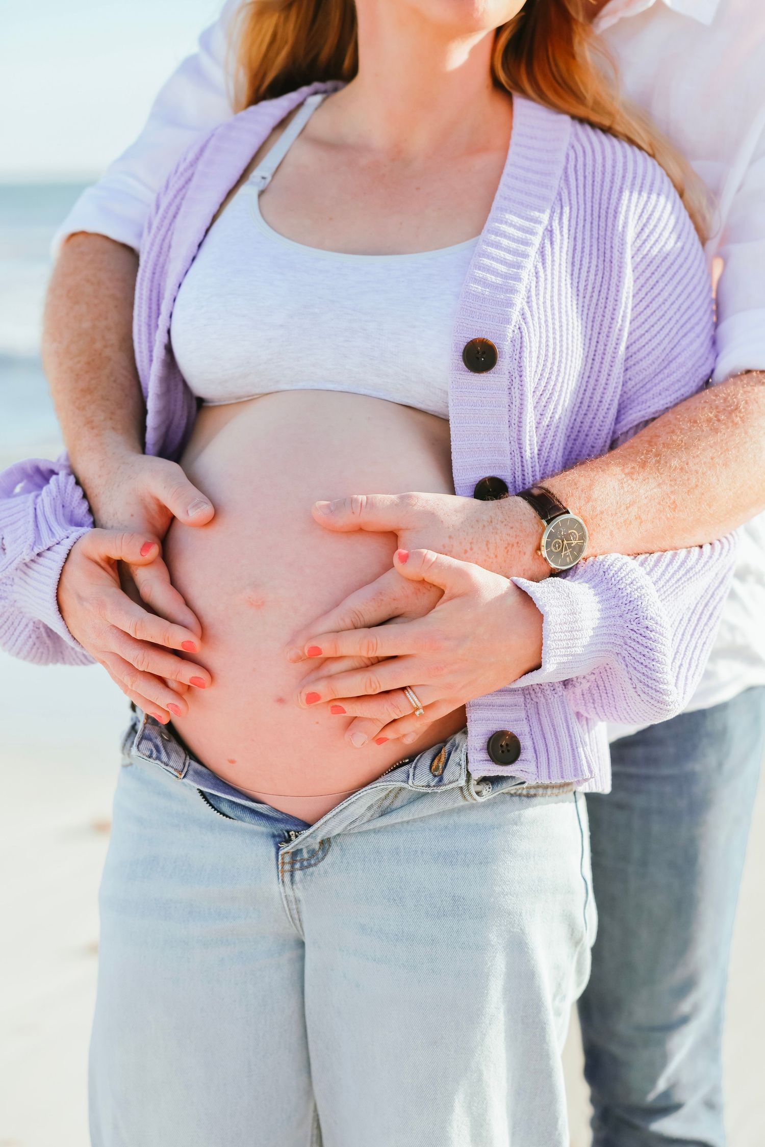 Couple holding pregnant belly together on beach, man wearing a watch and wedding ring, woman in purple cardigan.