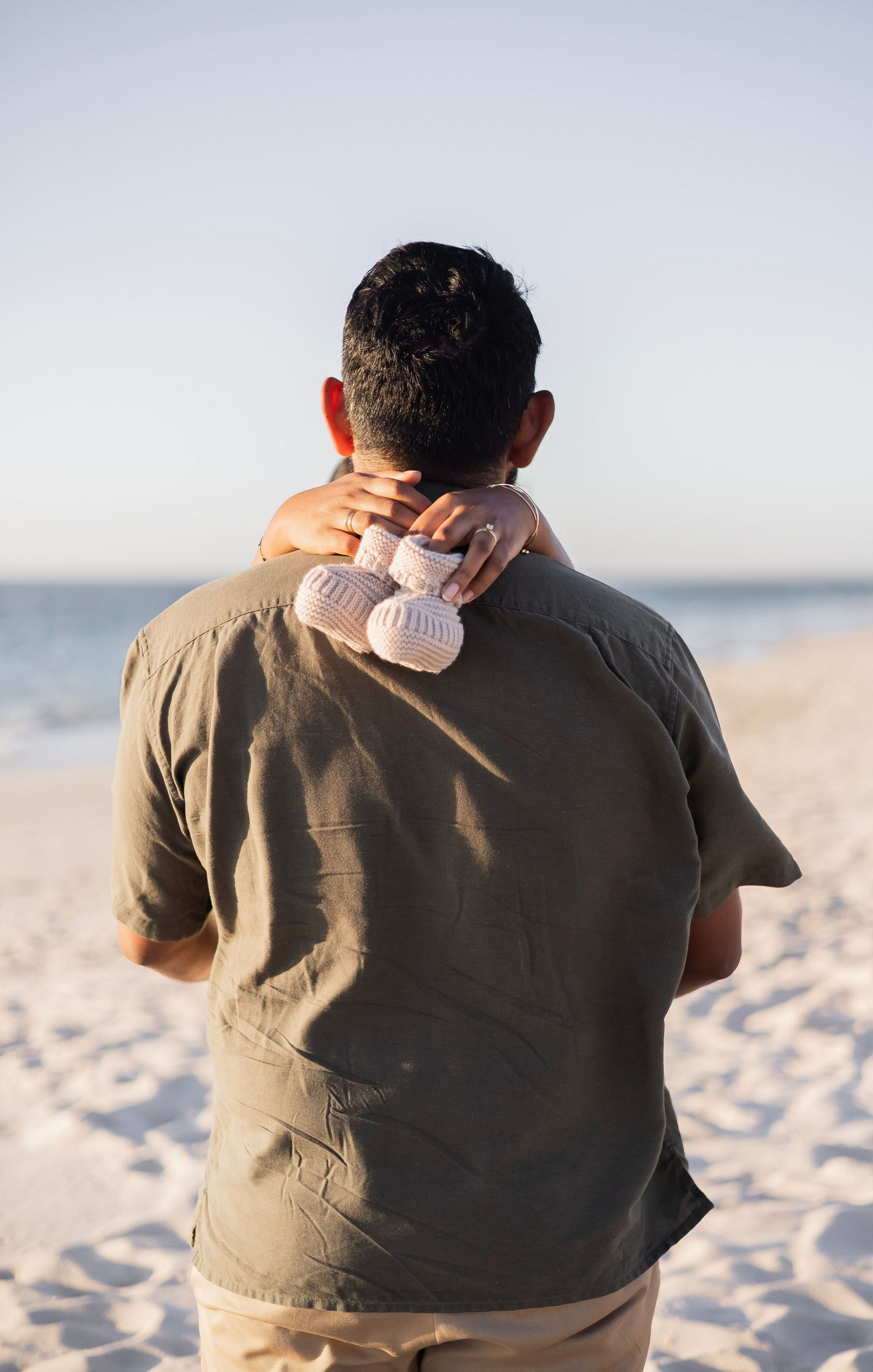 Man standing on a sandy beach holding pink baby shoes behind his back, viewed from behind.