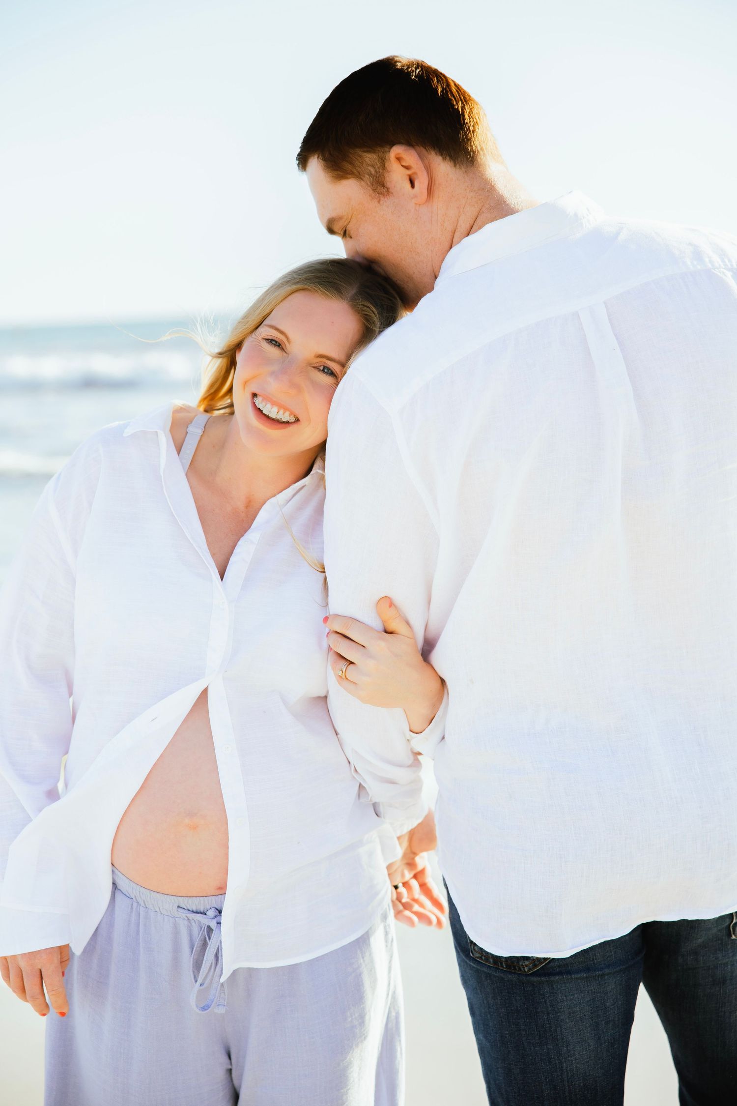 Smiling pregnant woman leans into partner's embrace on a sunny beach setting.
