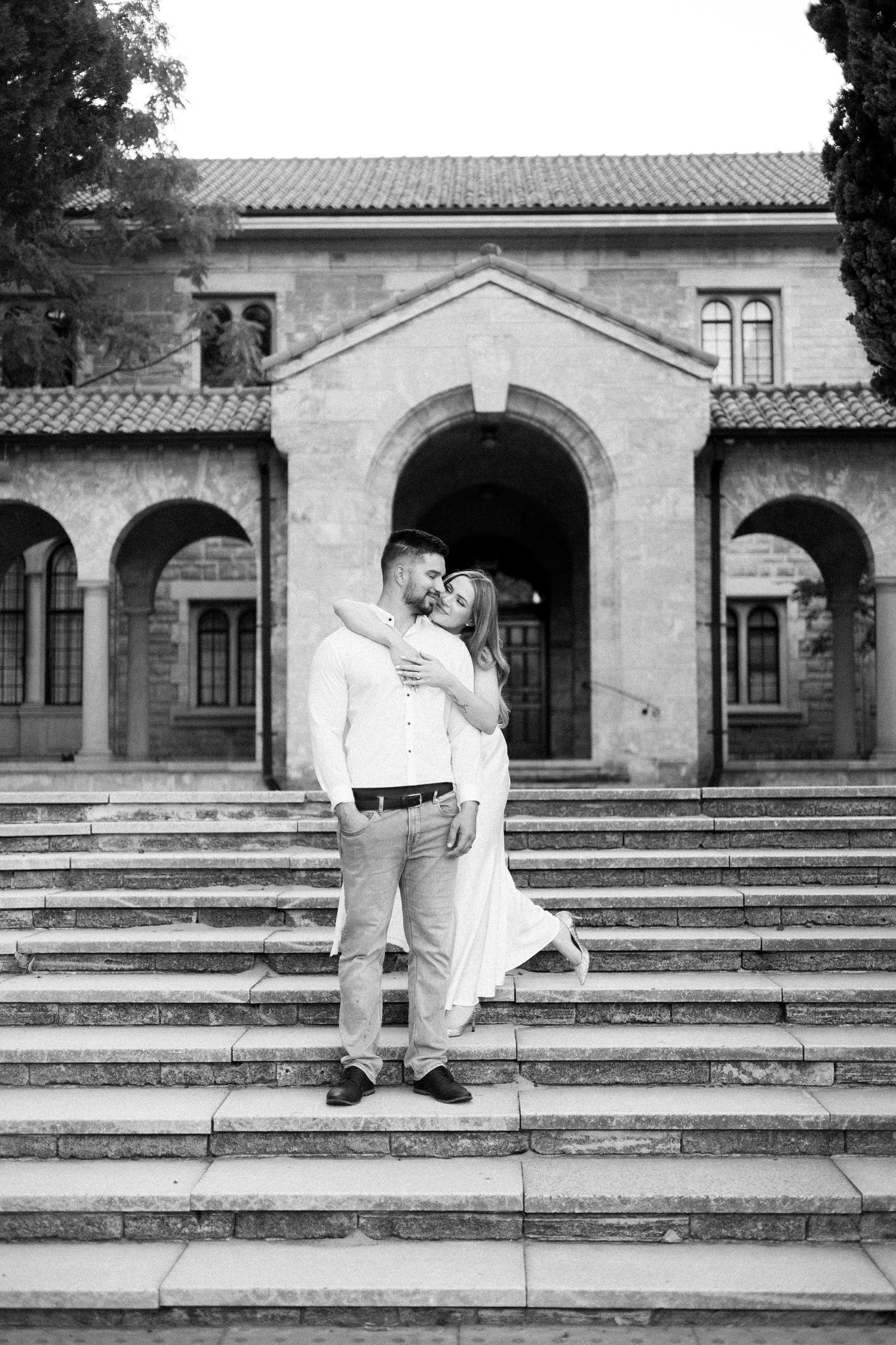 Black and white photo of couple embracing on wide stone steps in front of a historic arched building.