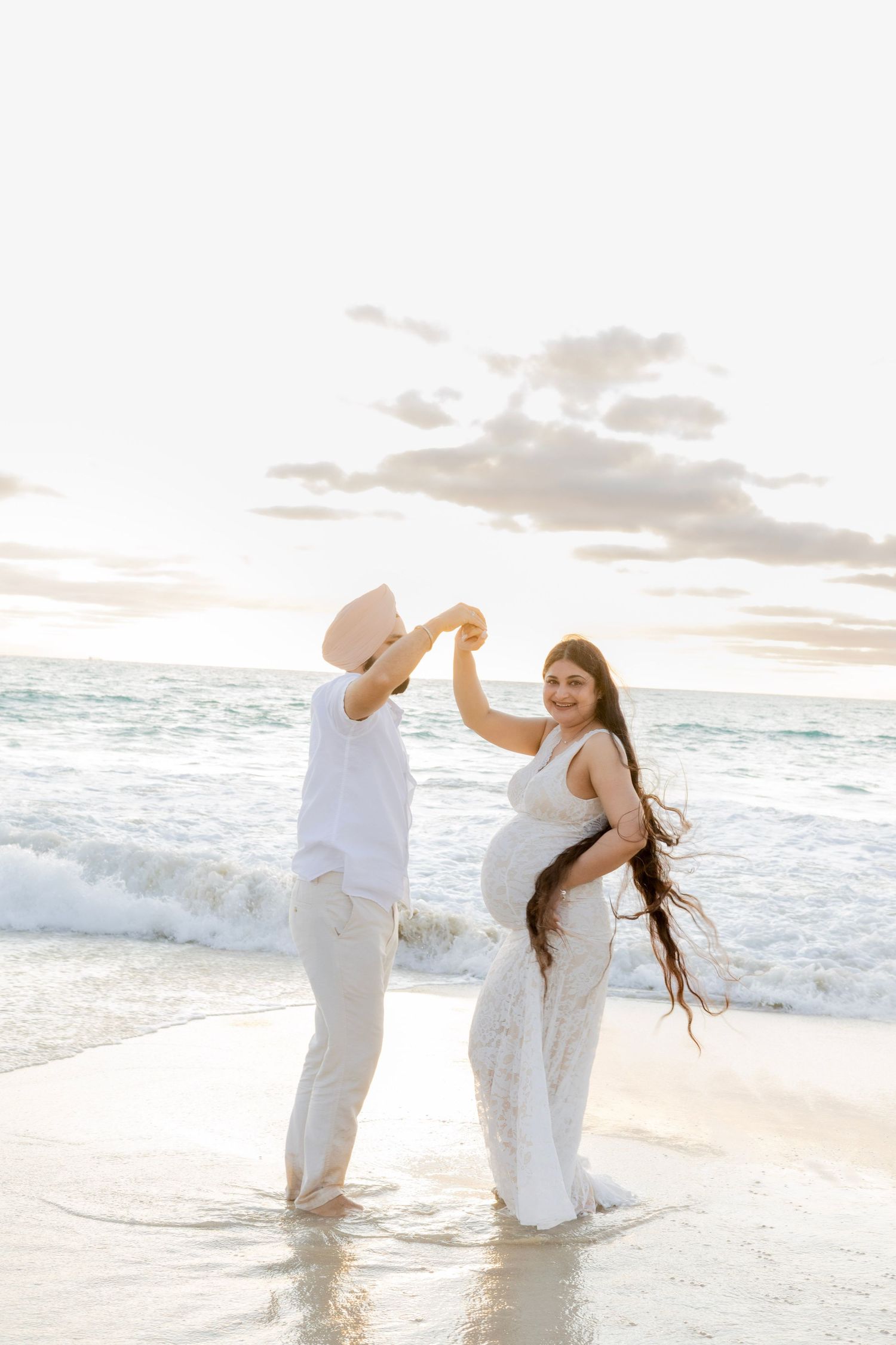 Pregnant woman in white lace gown dancing with partner in hat at beach sunset.