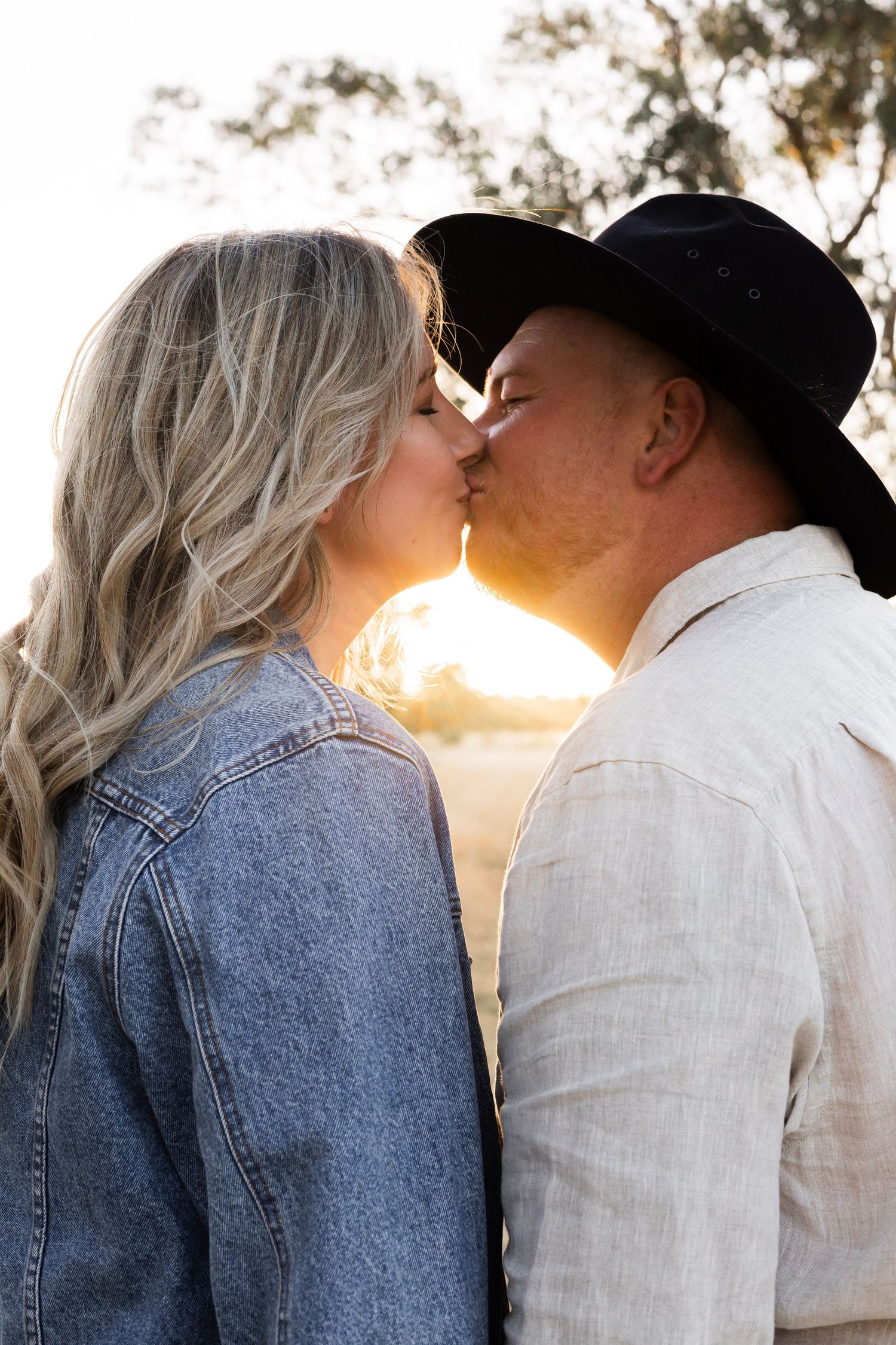 Couple sharing a romantic kiss backlit by a warm golden sunset in an open field.