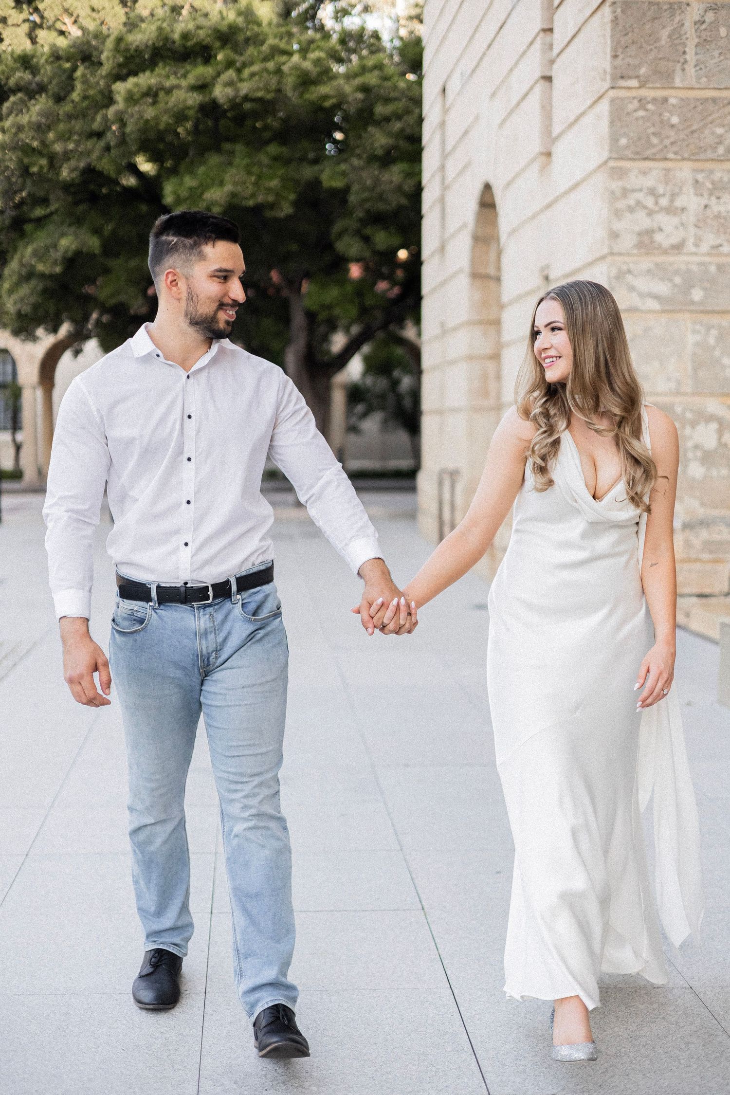 Smiling engaged couple holding hands walking past stone building, woman in white satin dress, man in jeans.