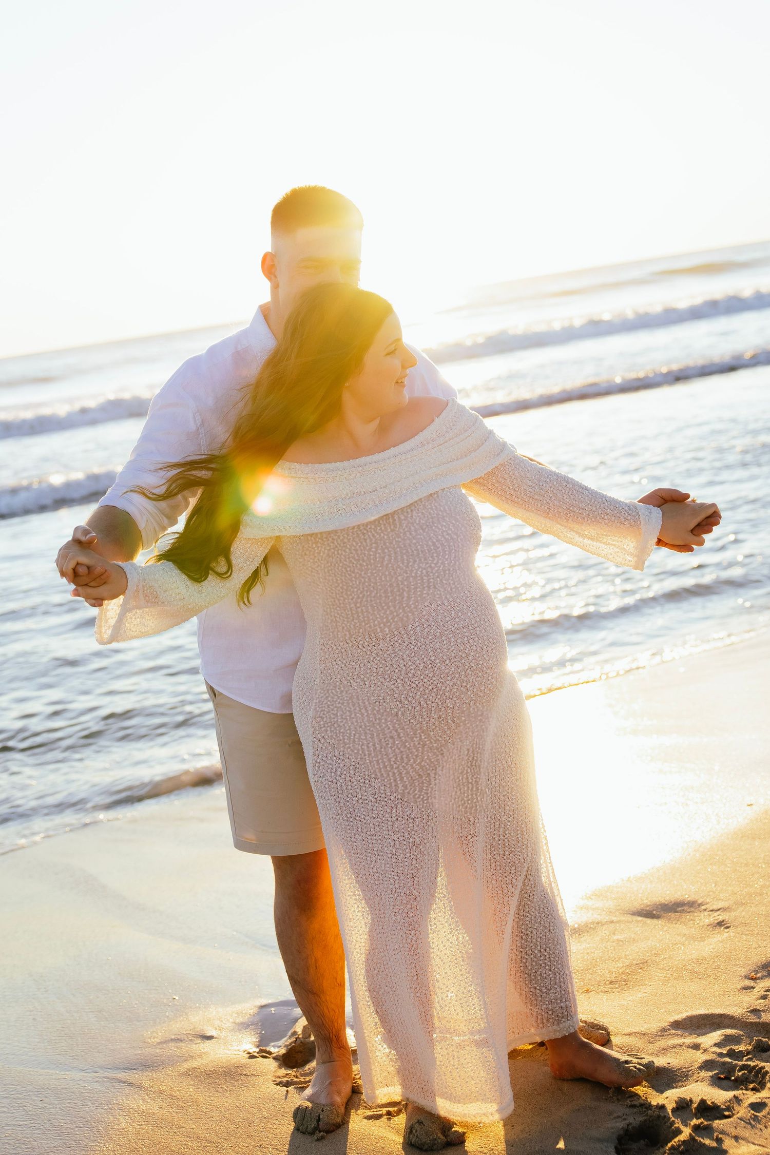Couple dancing romantically on a sandy beach at golden sunset, woman in white dress with arms outstretched.